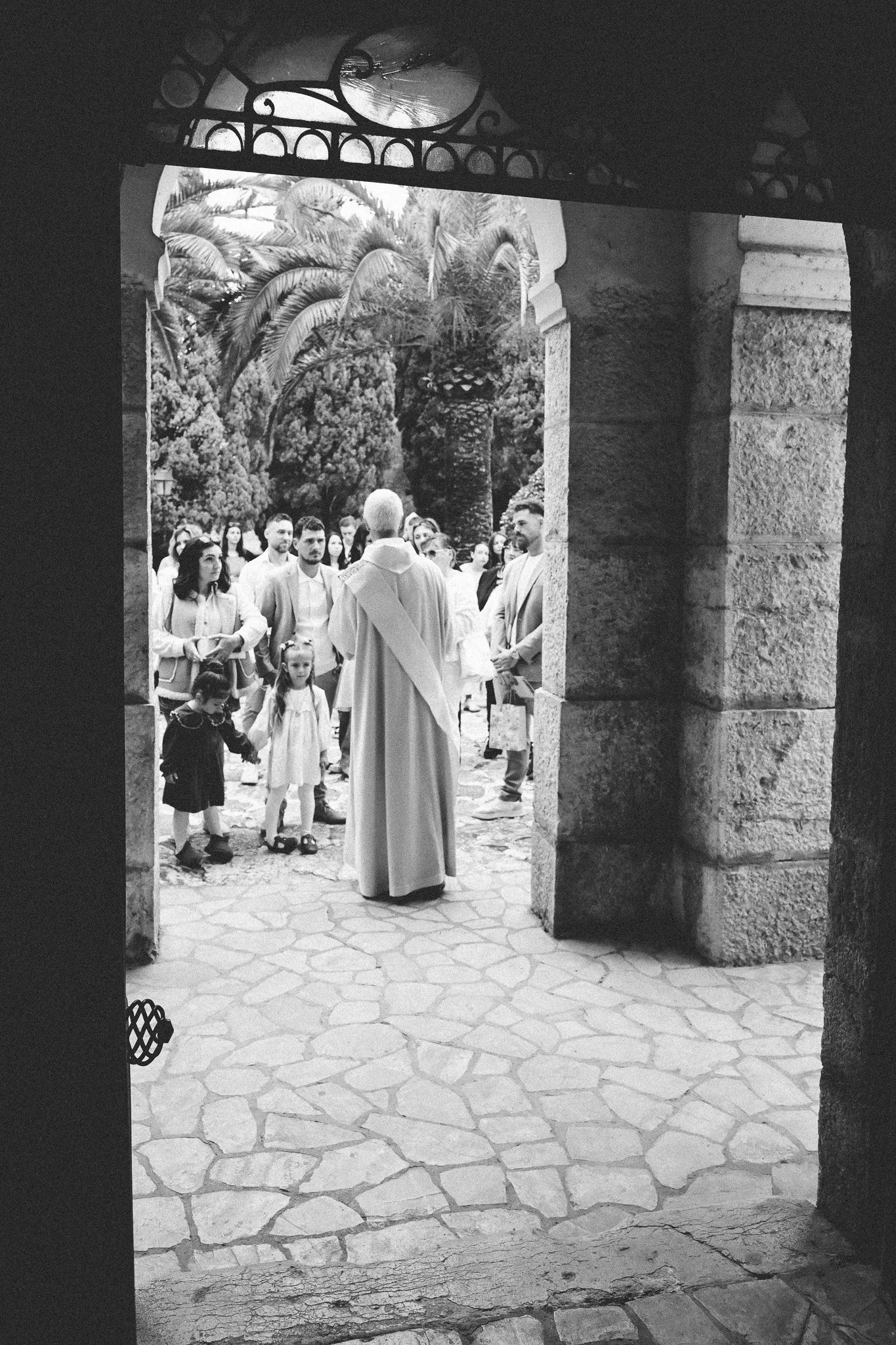 Une scène de groupe avec plusieurs personnes, incluant un enfant, vue à travers une arche de pierre. La scène se passe en extérieur avec des palmiers et des arbres en arrière-plan.