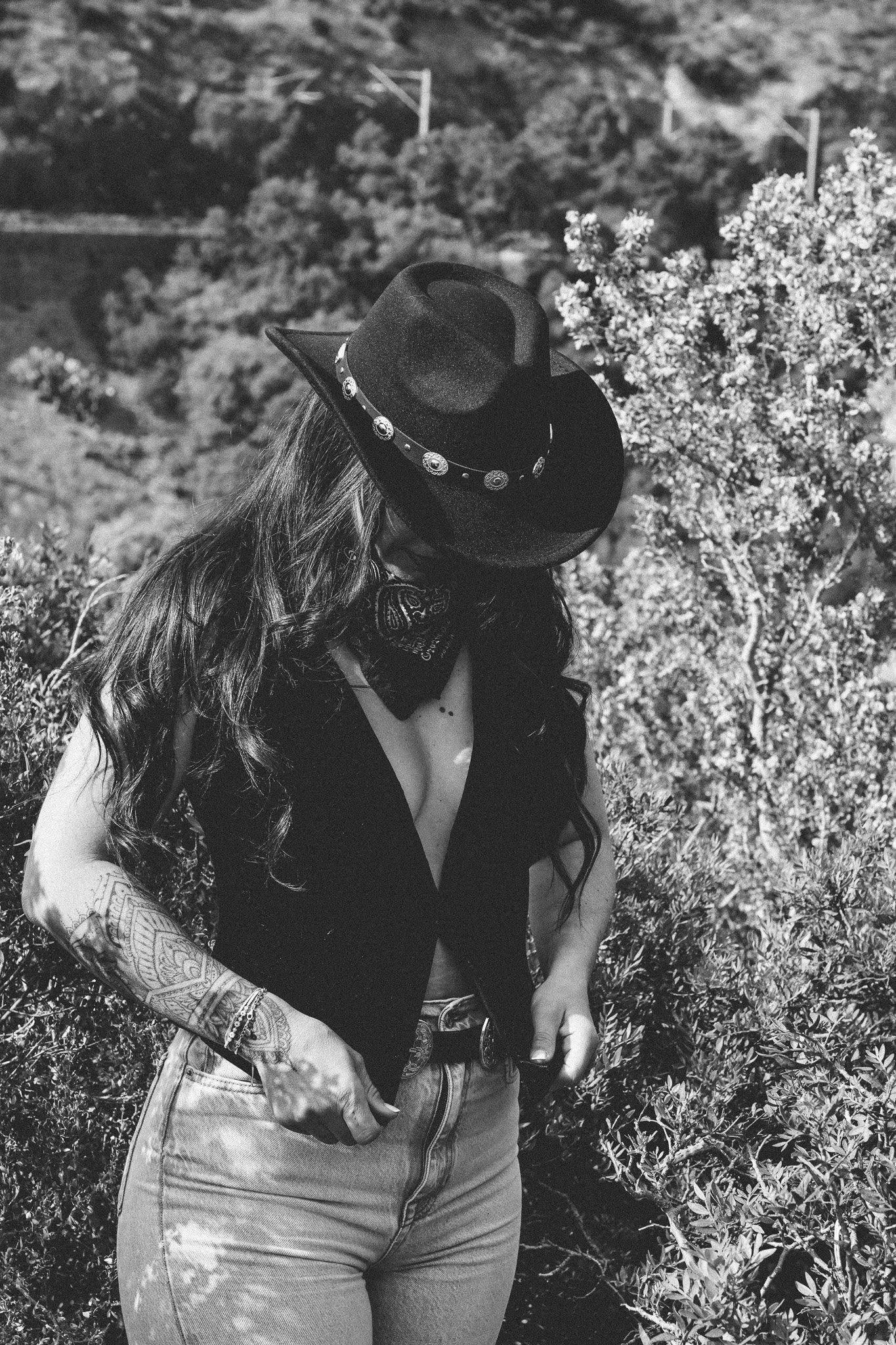 Une femme portant un chapeau noir avec des décorations, une veste sans manches, un bandana noir, des jeans et des tatouages sur le bras, se tient en extérieur parmi des buissons, vue en gros plan en noir et blanc.
