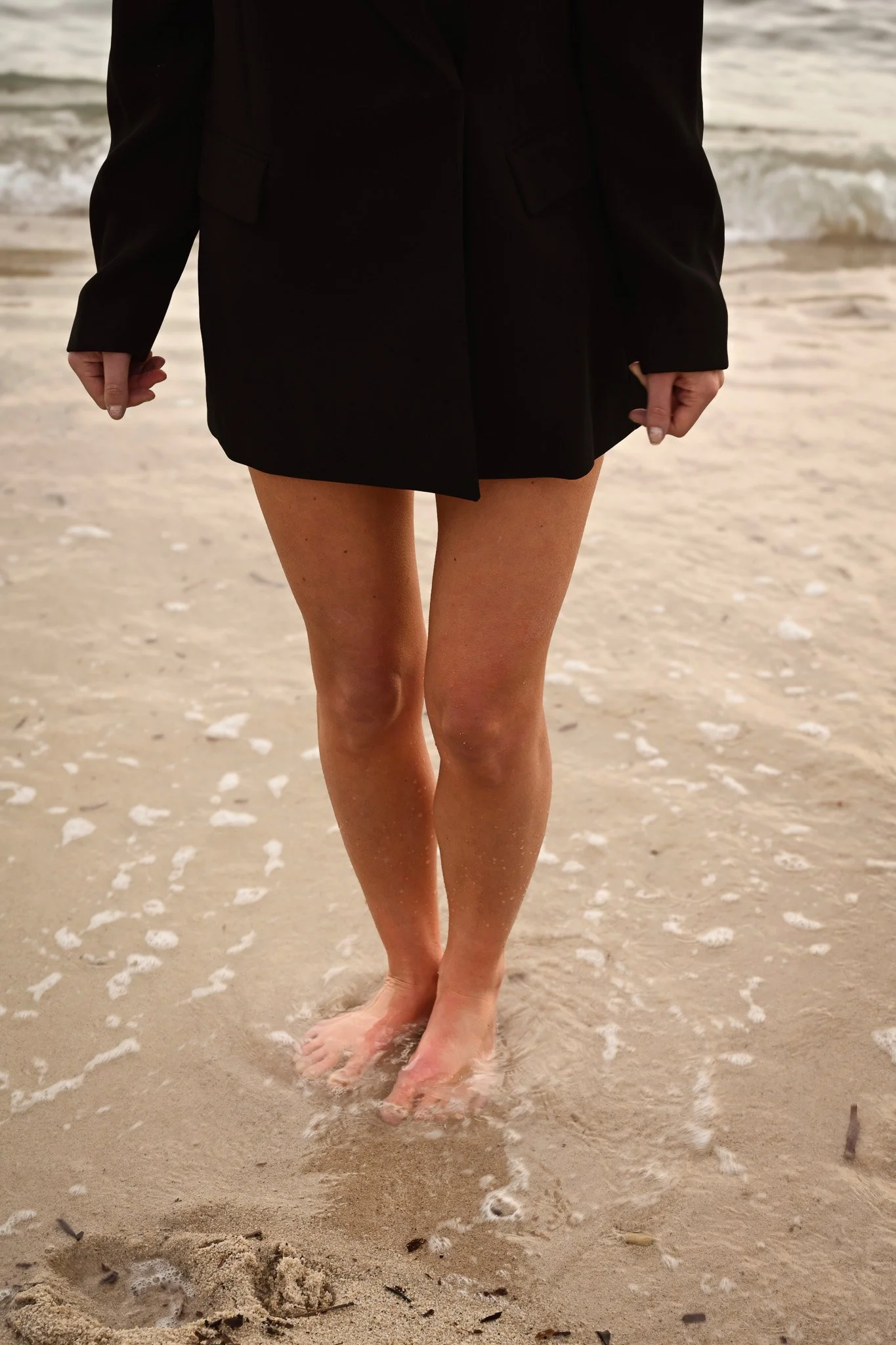 Une personne en veste noire marche pieds nus sur la plage, les vagues de l'océan derrière elle.