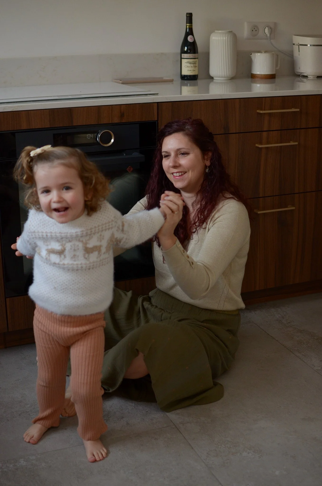 Une femme et une petite fille souriantes, se tenant la main dans une cuisine avec des meubles en bois, un four, une bouteille de vin, des vases blanc, et un appareil électroménager sur le comptoir.