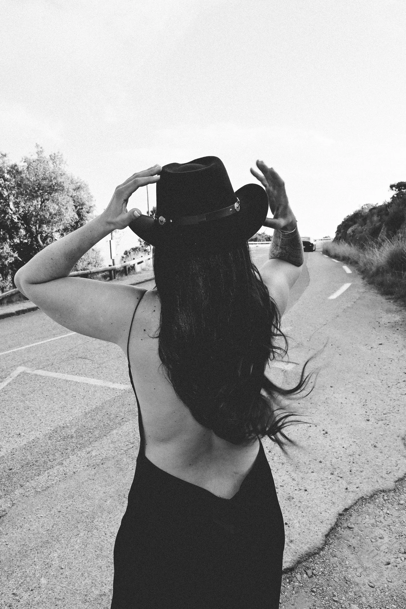 Femme avec longue chevelure porte un chapeau noir et pose le dos. Elle se tient au bord d'une route en plein air, avec des arbres à l'arrière-plan, en regardant au loin.