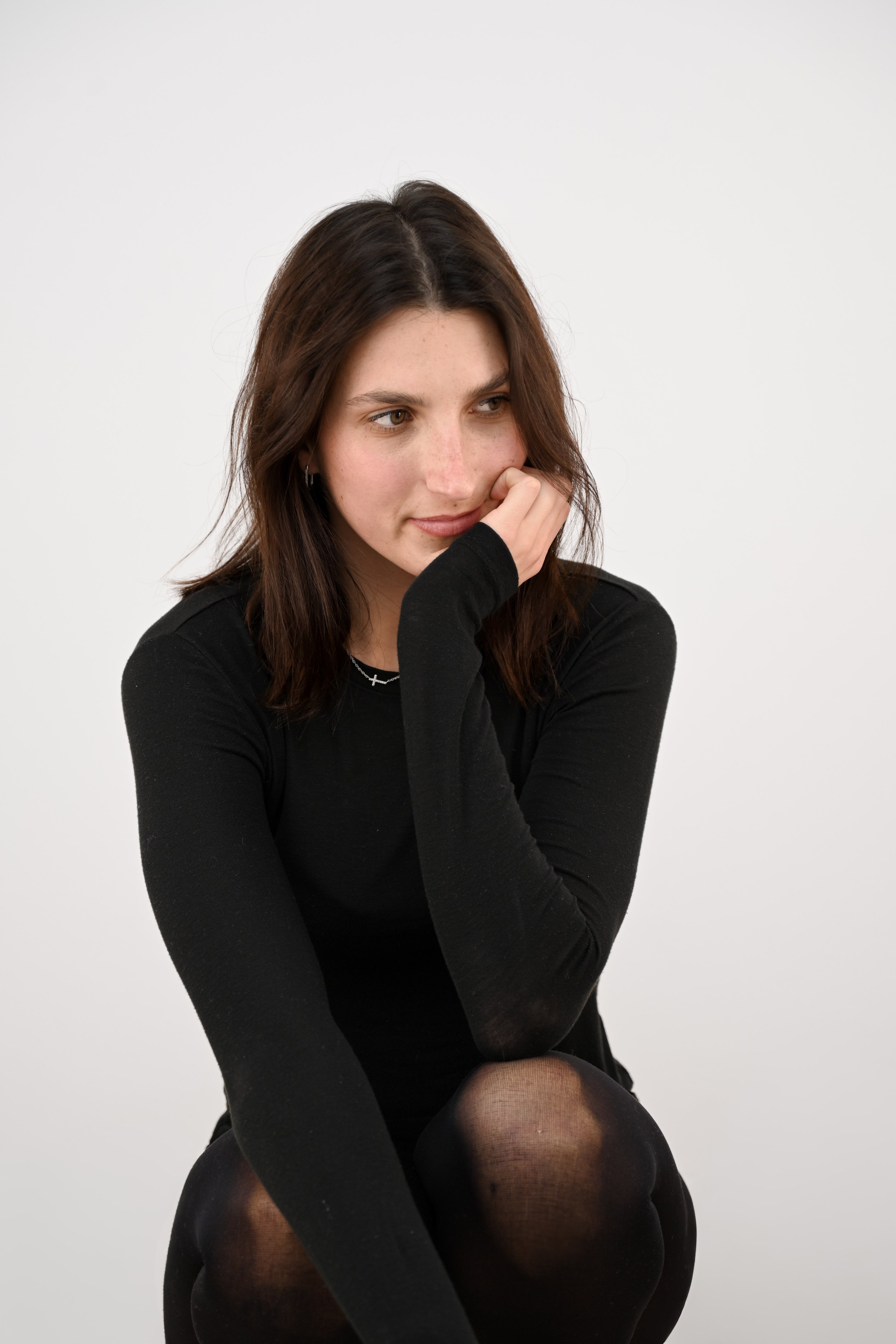 Une jeune femme aux cheveux bruns, habillée en noir, assise avec une main sur le visage, en regardant légèrement sur la droite, portant un collier en croix, dans un fond blanc.