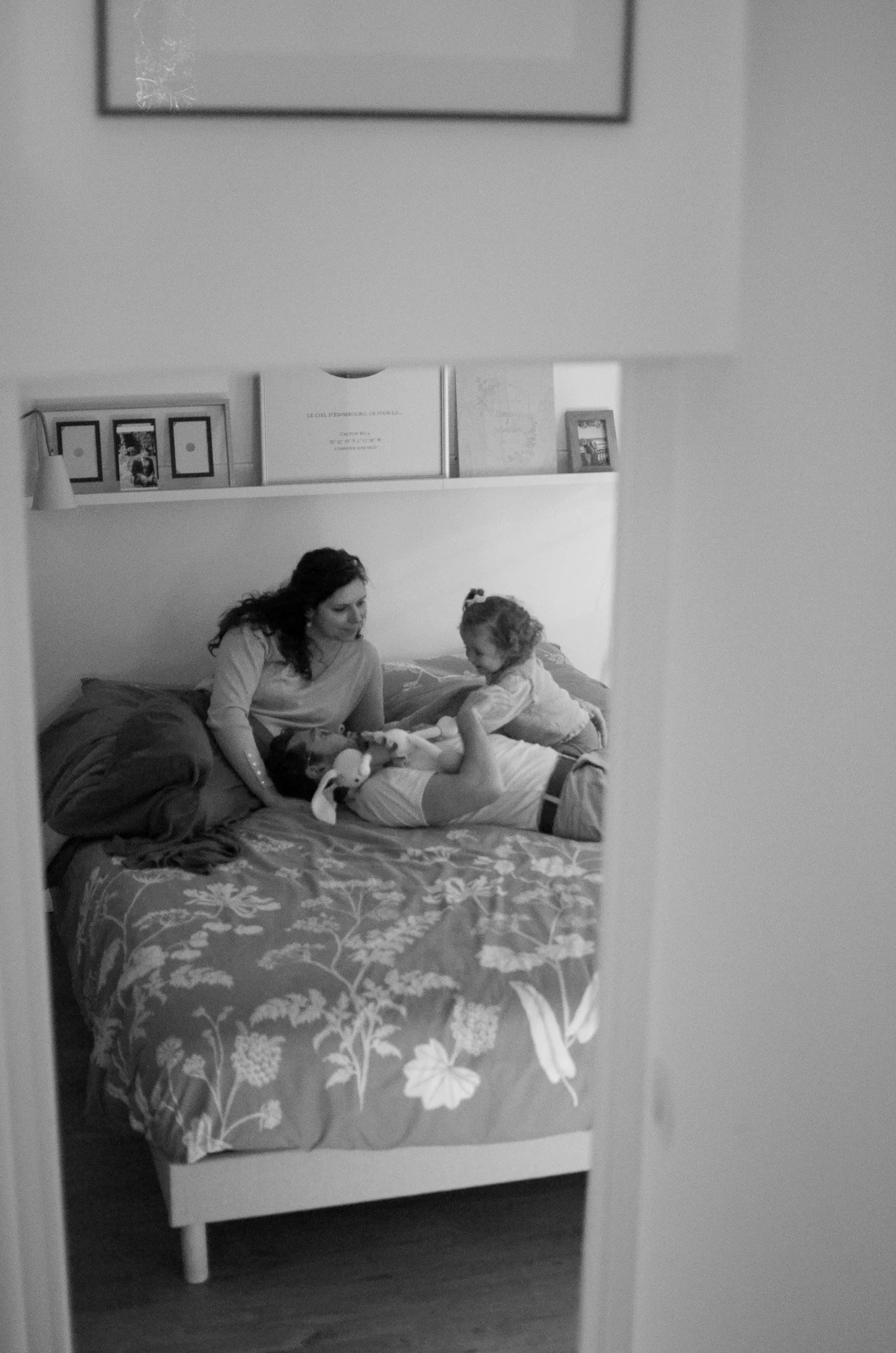 Une femme et un enfant jouent et se câlinent sur un lit dans une chambre, vue partielle à travers un miroir ou une porte ouverte.
