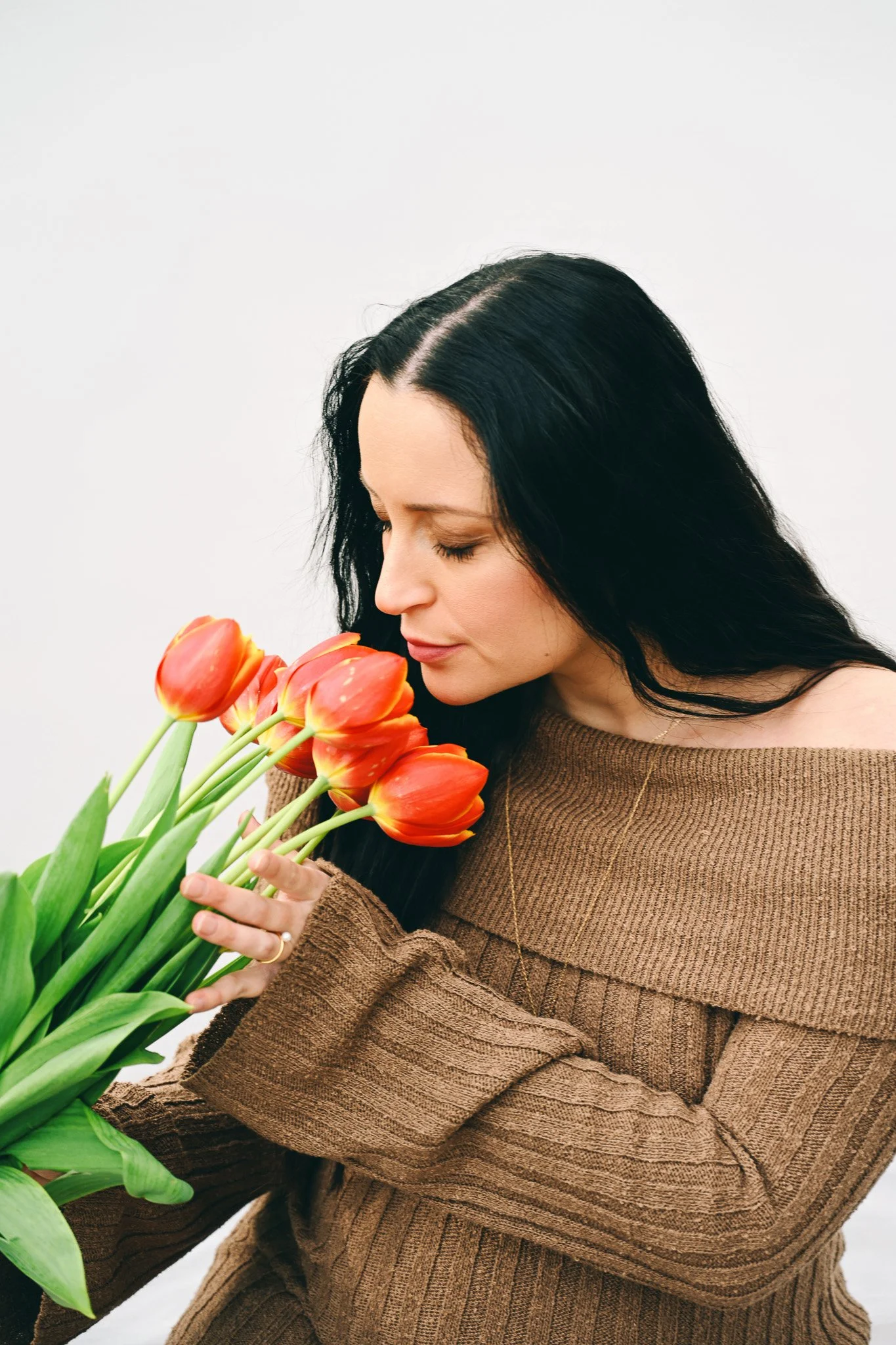 Une femme aux cheveux longs et noirs tient un bouquet de tulipes orange et rouges contre sa poitrine, vêtue d'un pull marron.