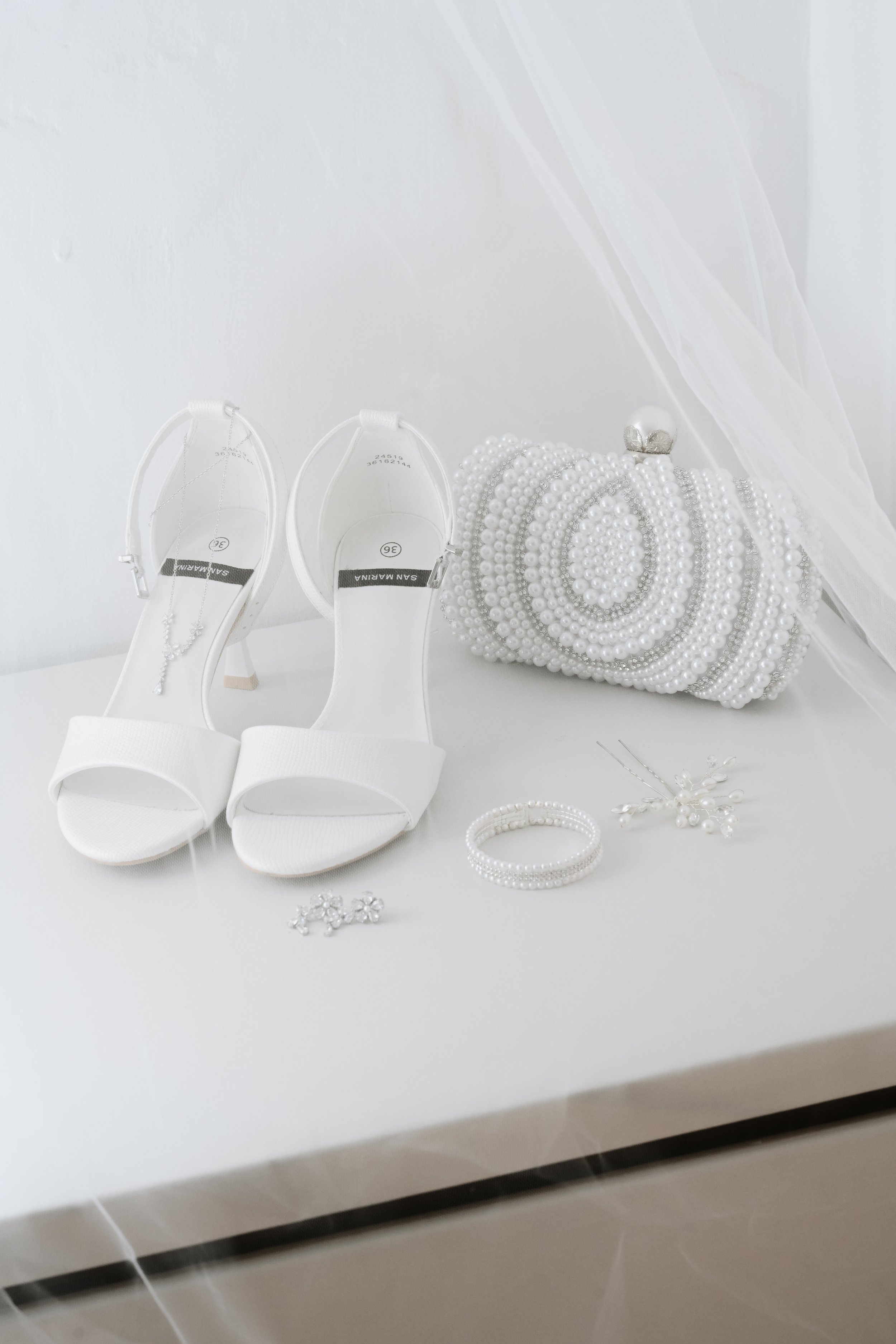 Chaussures blanches à talons, accessoires de bijoux blancs, perles, bracelet, broche et boucles d'oreilles posés sur une table blanche.