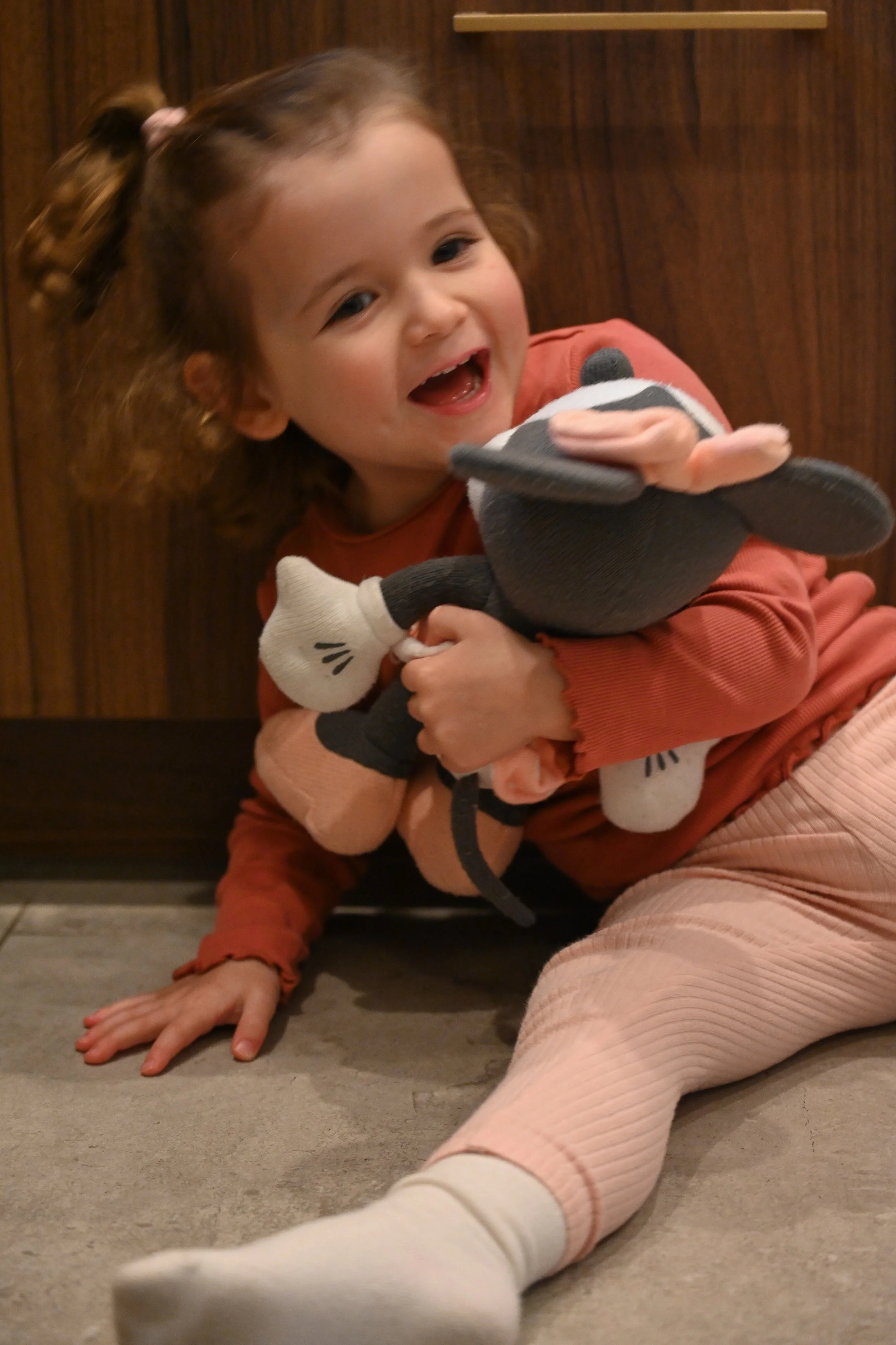 Une petite fille souriante avec des cheveux roux noués en deux avec des rubans, portant un pull orange et des leggings rose clair, qui tient un pantoufle en peluche d'animal noir et blanc.