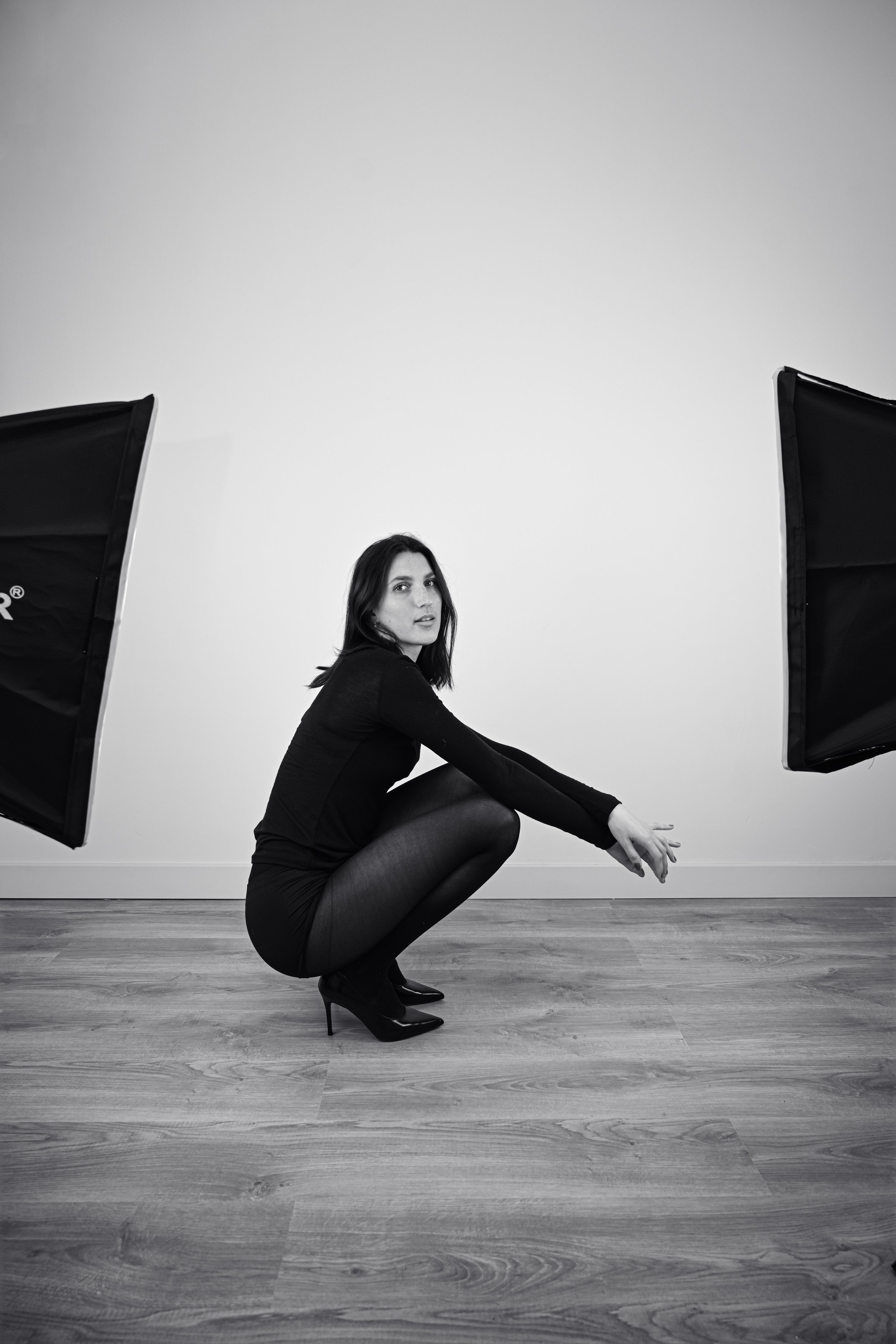 Une femme en vêtements noirs et talons aiguilles noirs, assise en pliant les jambes, dans une pièce avec un sol en bois, deux réflecteurs de studio sont placés de chaque côté derrière elle.