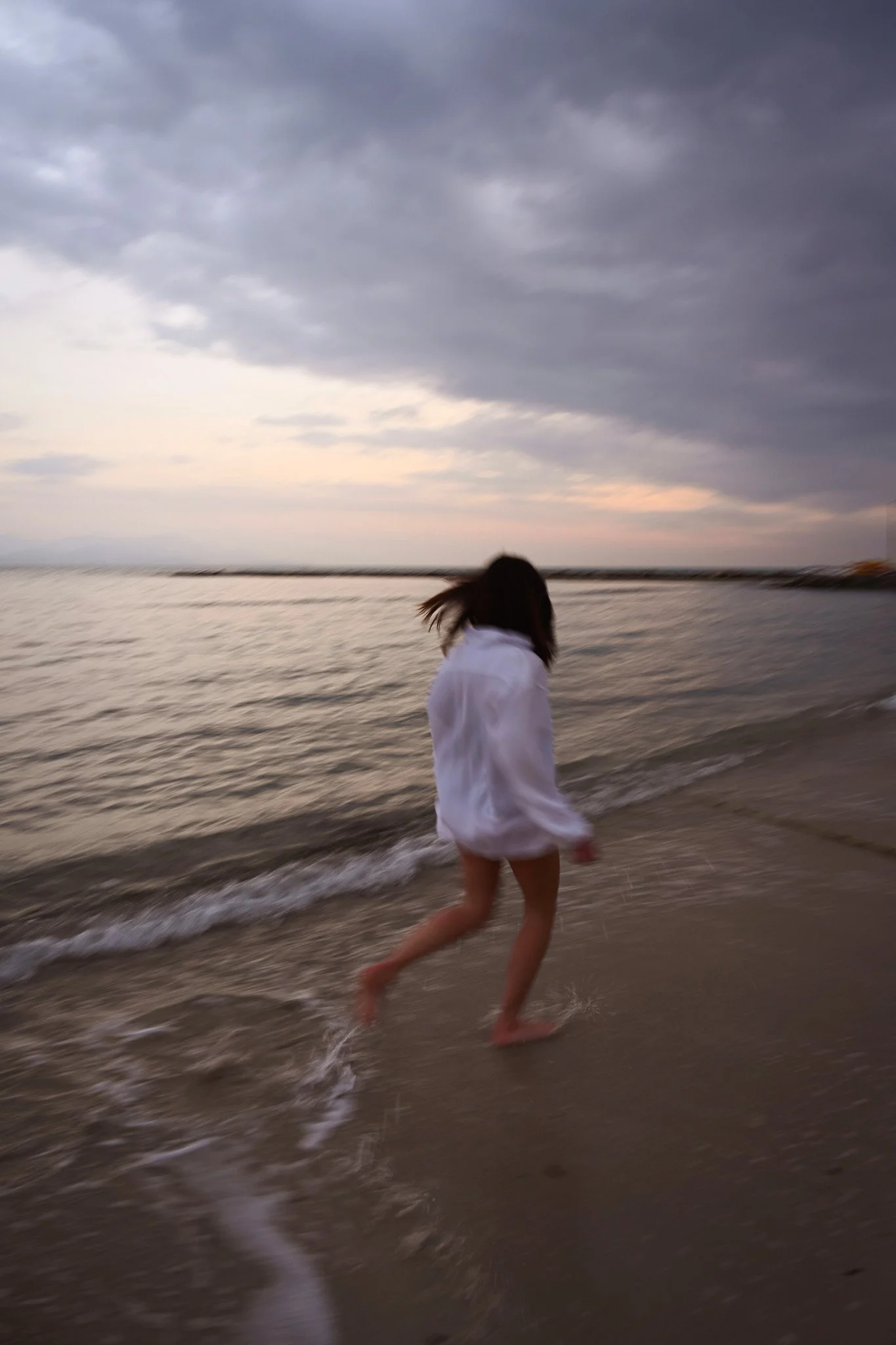 Une personne portant une robe blanche court sur la plage au bord de l'océan, avec un ciel nuageux en arrière-plan, probablement au coucher ou au lever du soleil.