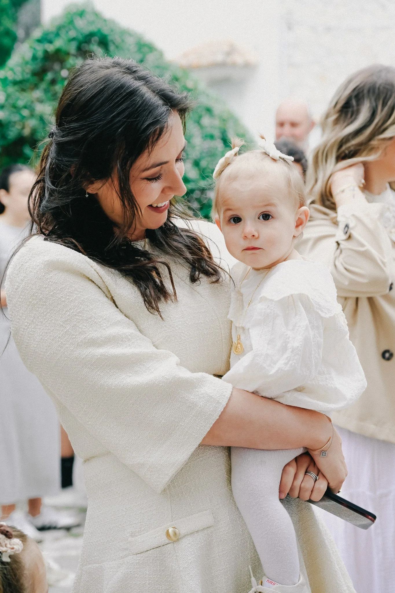 Une femme porte une petite fille blanche lors d'un événement civil ou religieux, avec d'autres personnes floues en arrière-plan.