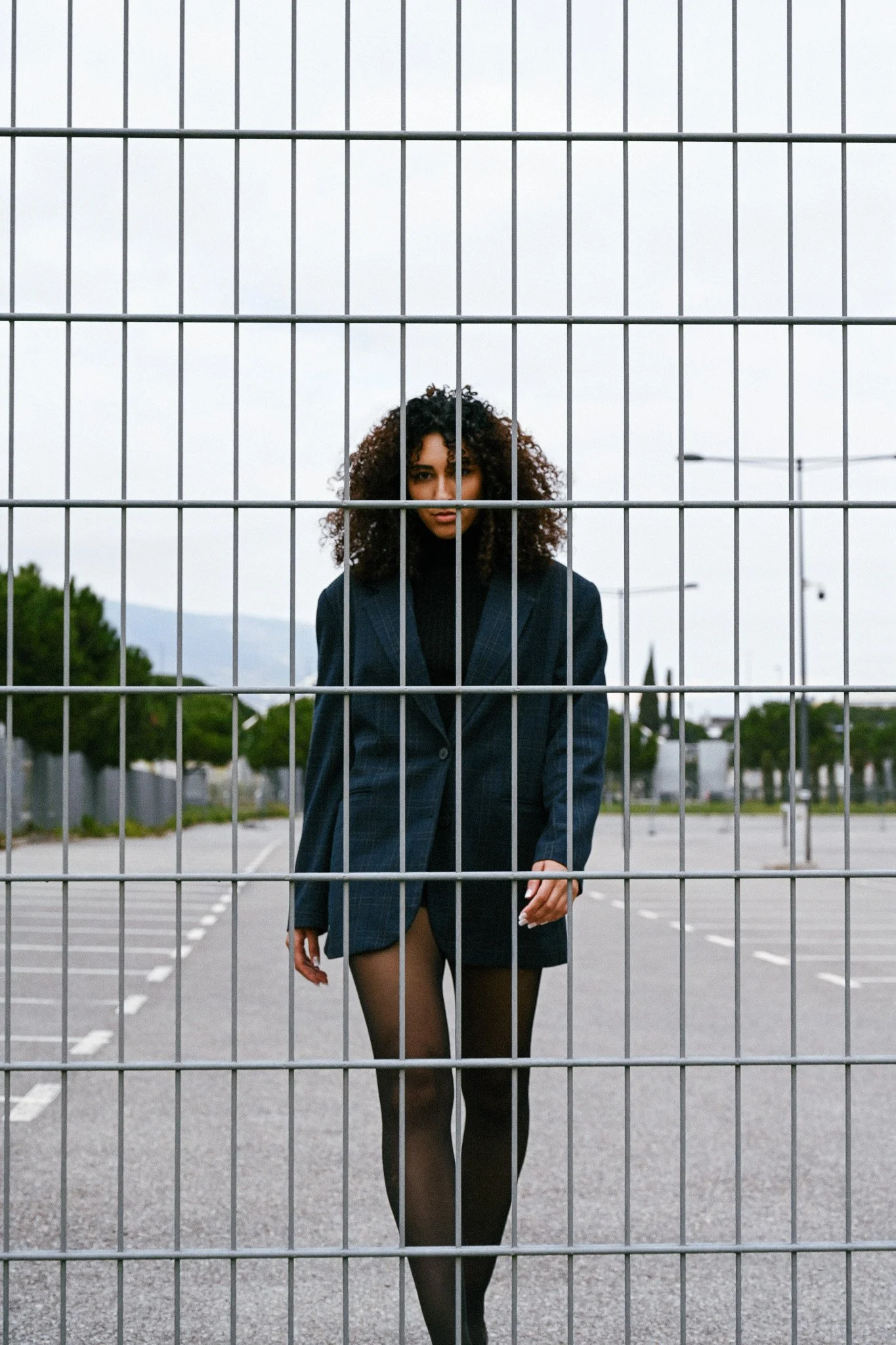 Une femme en tenue sombre, avec des collants noirs, se tient derrière une barrière métallique dans un parking ou un terrain de sport. Elle regarde vers l'objectif avec un sourire confiant.