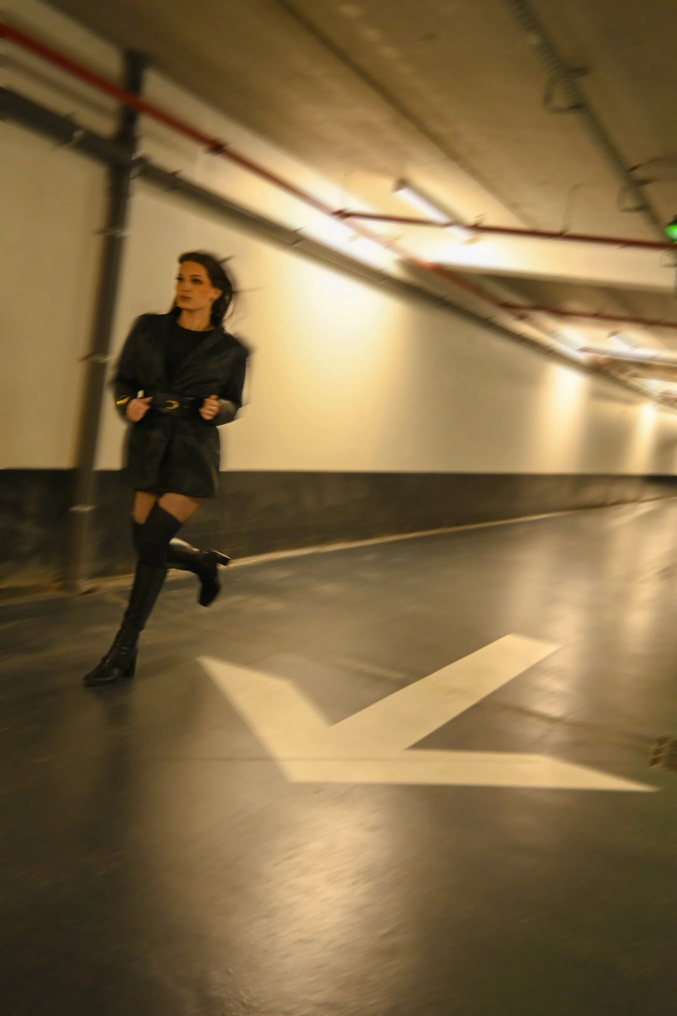 Femme marchant dans un corridor souterrain en noir, portant des bottes hautes, avec des tuyaux au plafond.