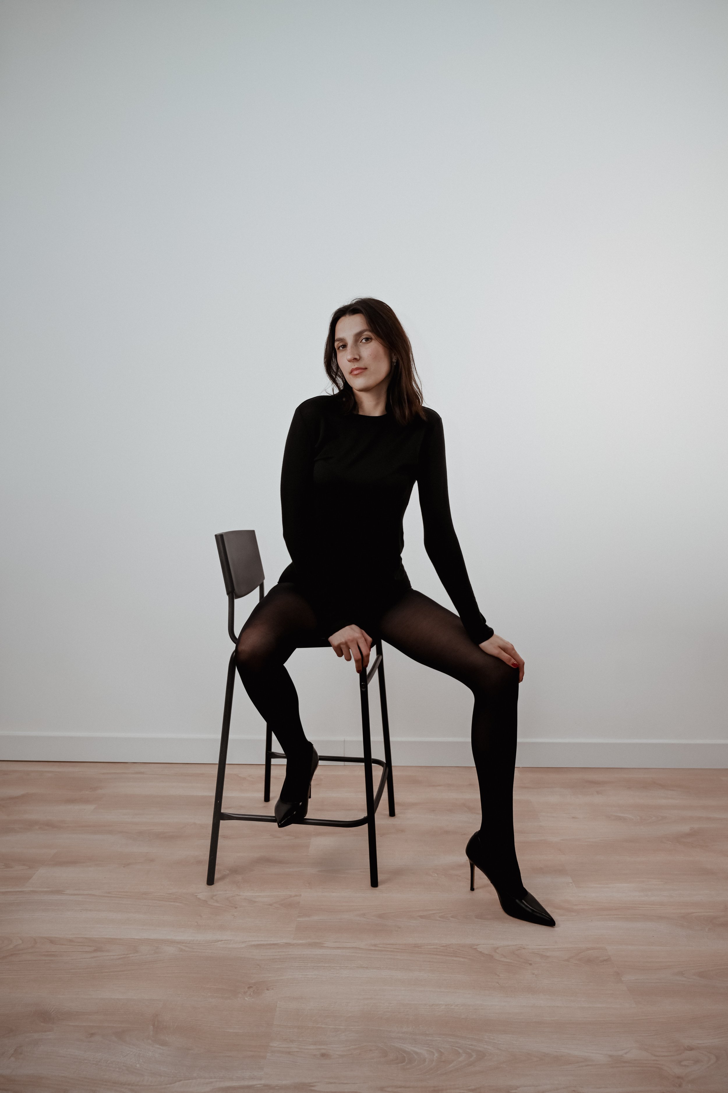 Une femme avec des cheveux bruns, portant une robe noire, assise sur un tabouret en bois avec une seule jambe sur le sol et l'autre sur un tabouret, dans une pièce avec un mur blanc et un plancher en bois clair.