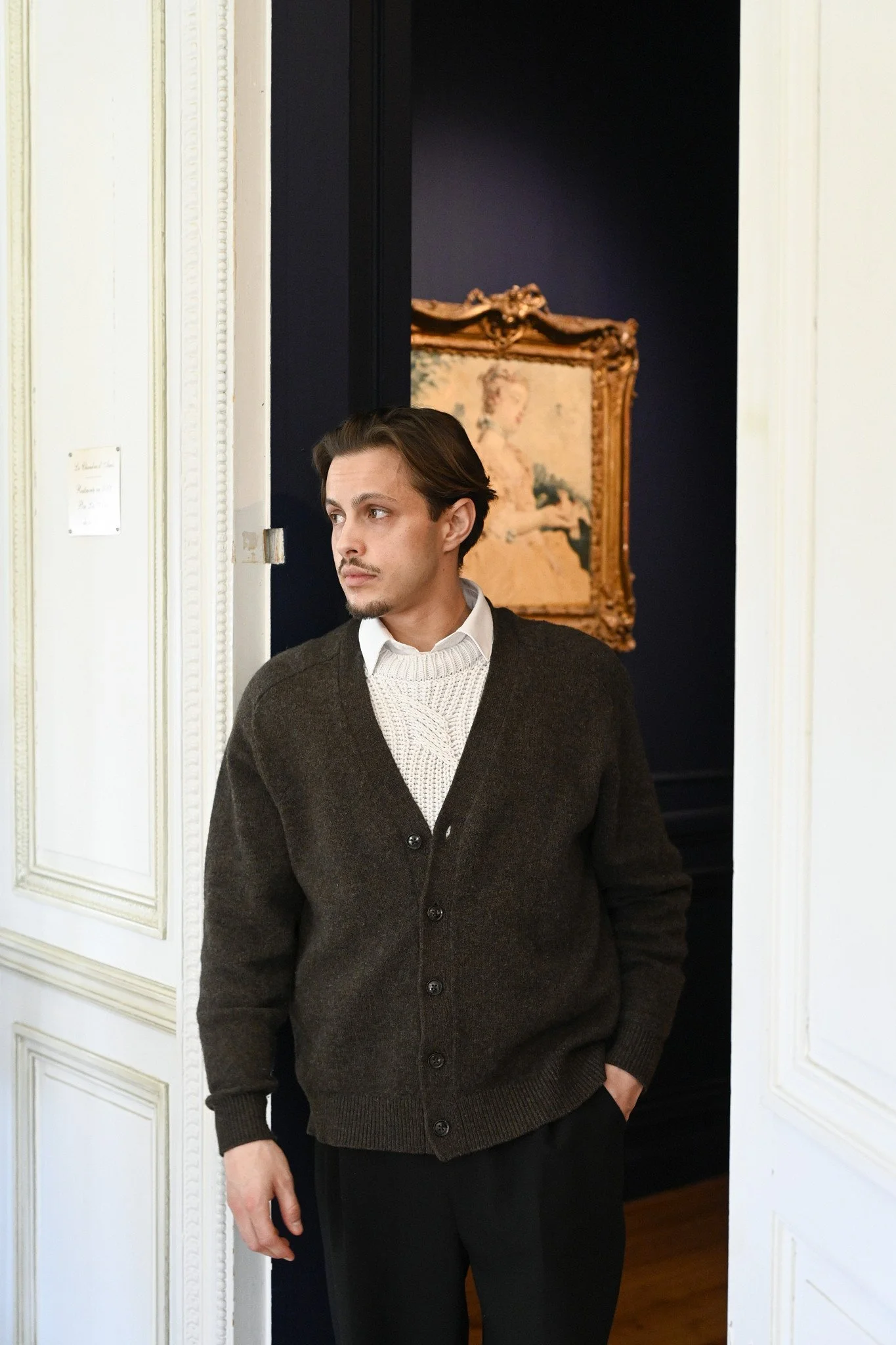 Un homme dans un veston brun foncé et un pull blanc, debout dans une pièce à l'intérieur, avec un portrait ancien en fond dans un cadre doré.