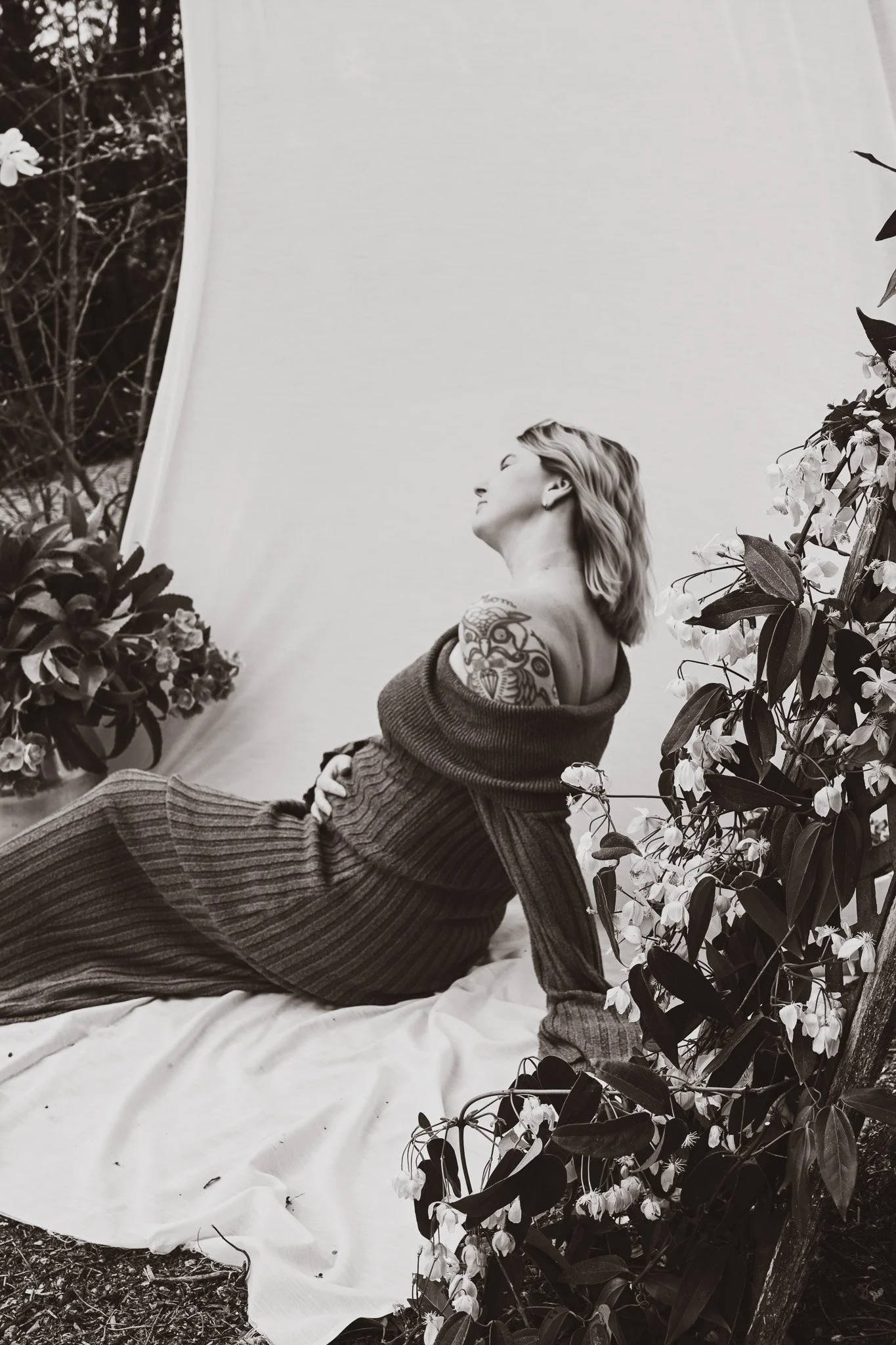 Une femme assise sur une couverture blanche entourée de plantes avec des fleurs, regardant vers le haut, en noir et blanc.