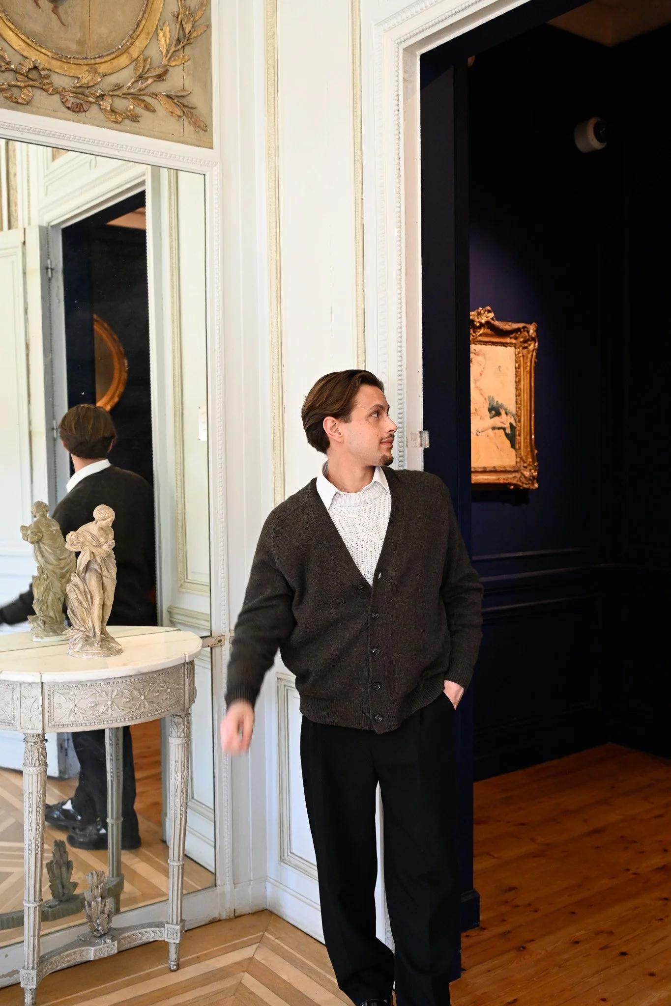 Un jeune homme en veste marron et pantalon noir se tient dans une salle d museum, à côté d’un miroir et d’une petite table avec des sculptures en marbre, en face d’un mur blanc avec des cadres dorés et une peinture dans un cadre doré.