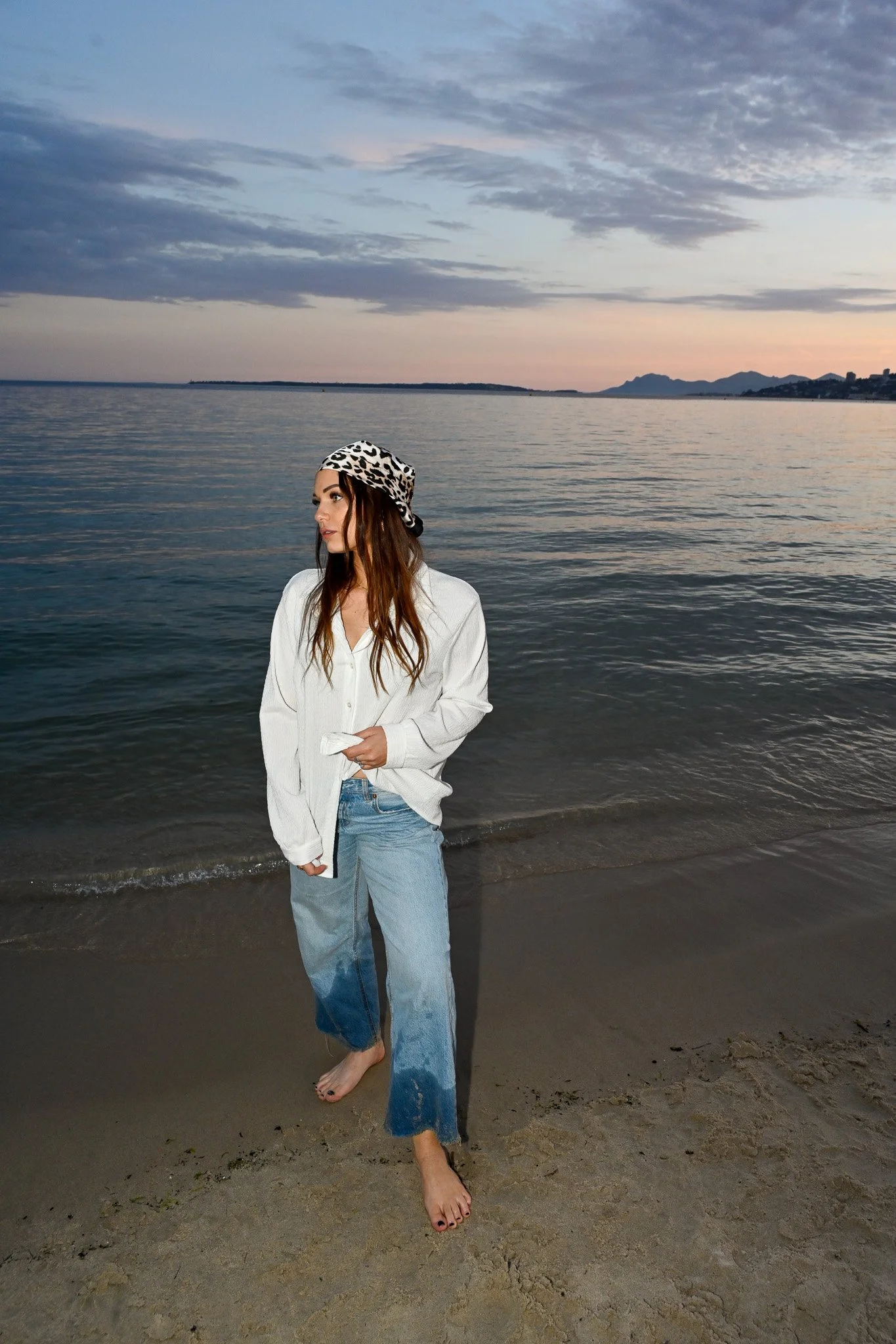 Jeune femme avec un chapeau léopard, portant un pantalon déchiré, pieds nus, se tenant sur la plage au bord de l'eau, au coucher ou lever du soleil avec des montagnes en arrière-plan.