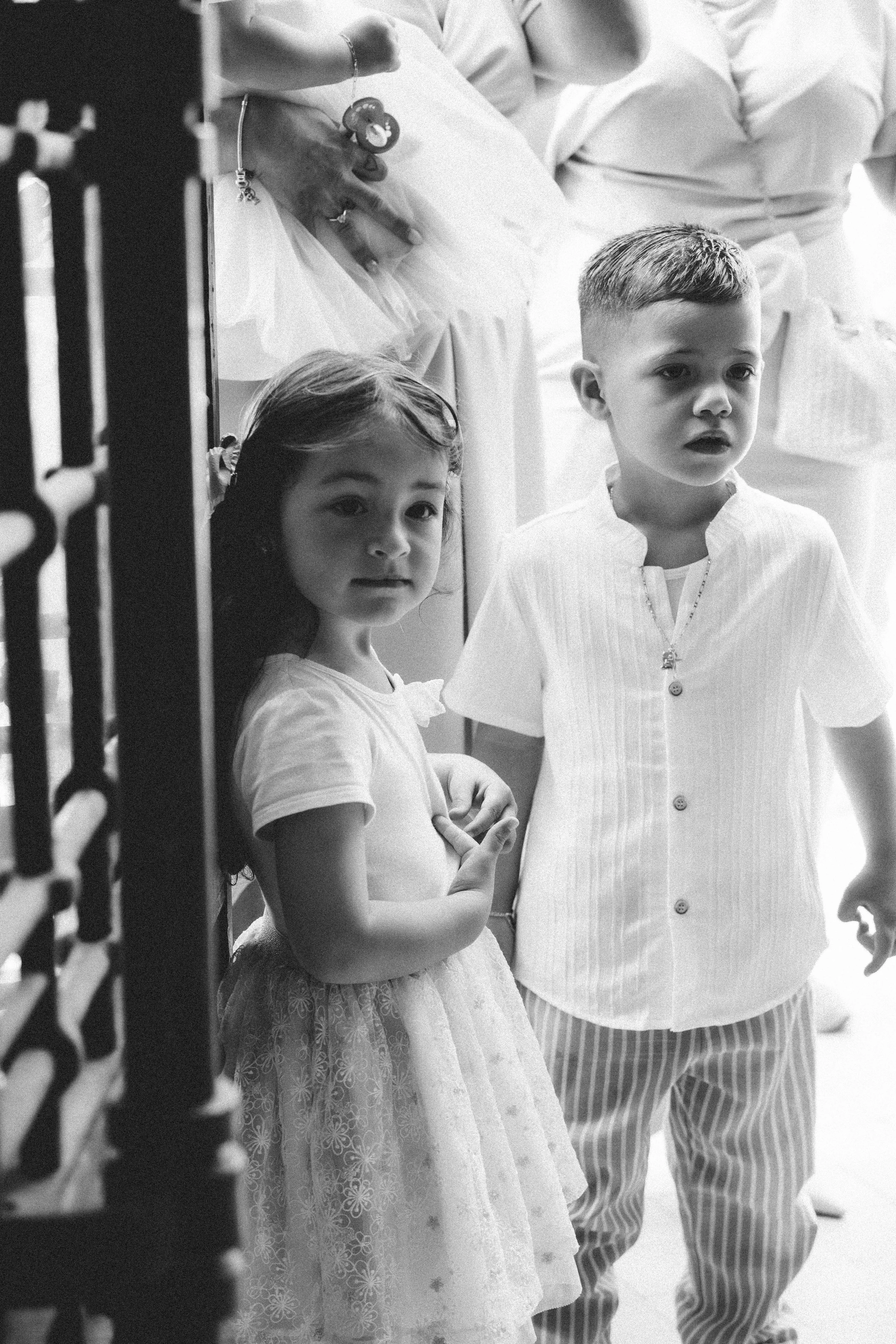 Deux enfants, une fille et un garçon, portent des vêtements légers et se tiennent côte à côte. La fille porte une robe en tulle avec des motifs floraux, et le garçon porte une chemise à rayures avec un pantalon assorti. Leur expression est neutre. Il y a une personne adulte derrière eux, tenant un objet, mais elle n'est pas entièrement visible.