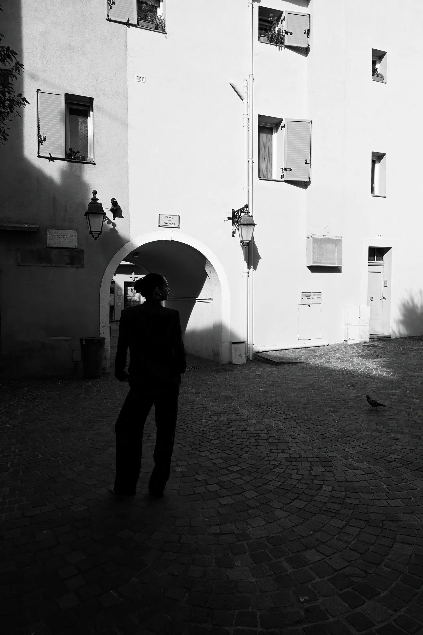 Une scène urbaine en noir et blanc avec une silhouette de femme regardant un pigeon sur un pavé. Il y a des fenêtres ouvertes, des lampes anciennes, et un mur avec un panneau indiquant « Place du Couvent ». Un arc forme un passage sous un bâtiment, e