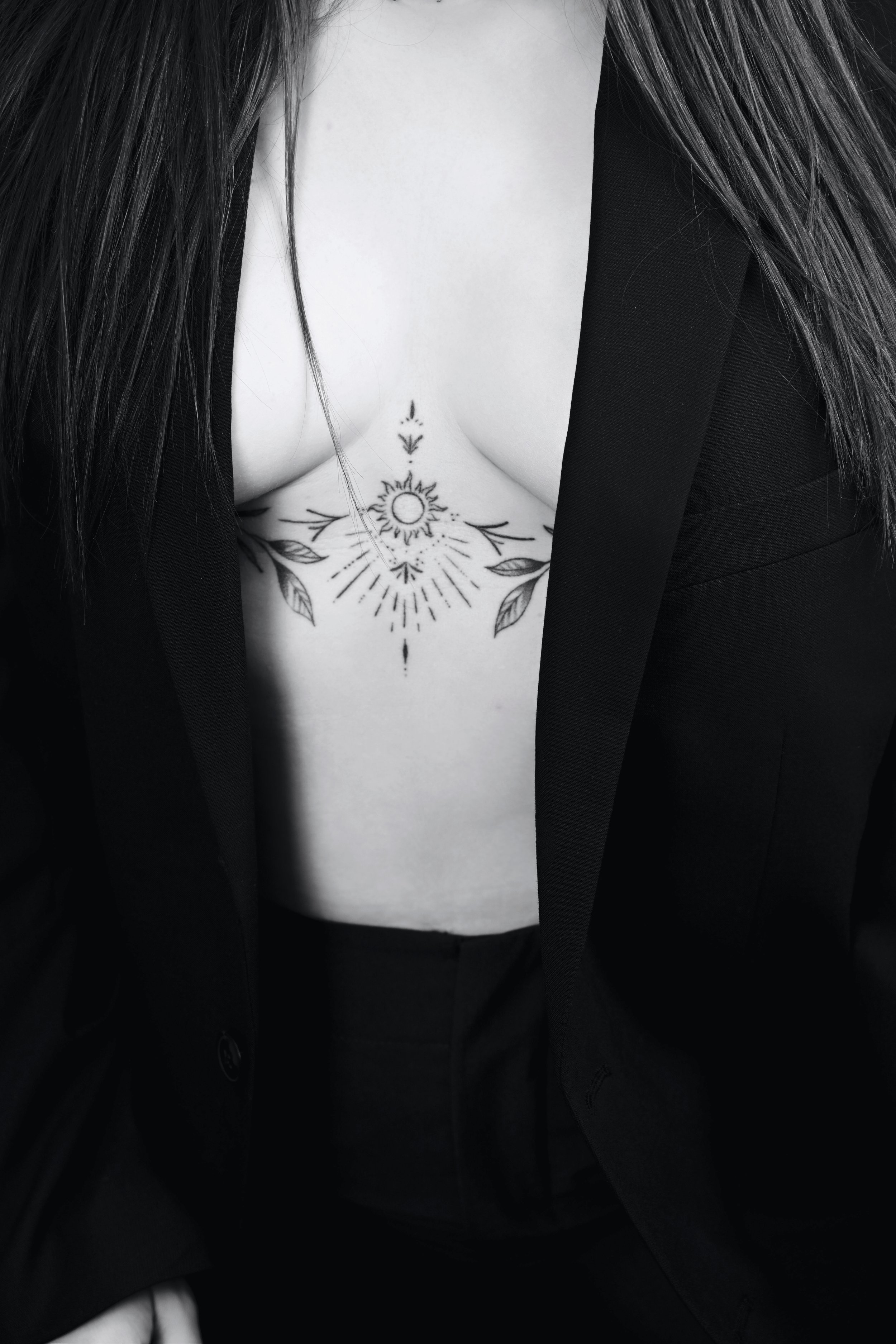 Une femme porte une veste noire avec un décolleté révélant un tatouage de soleil et de lignes géométriques sur la poitrine.