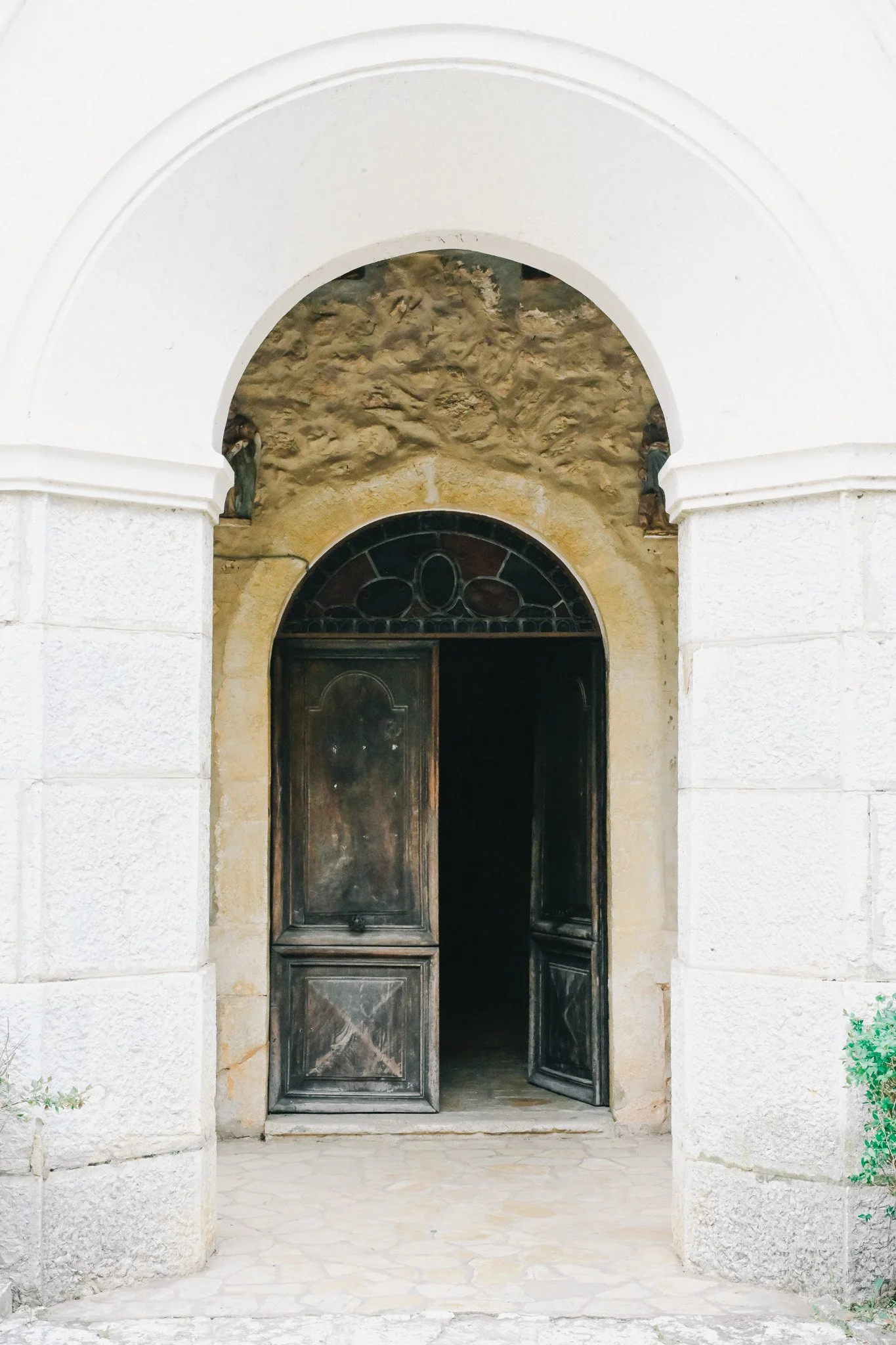 Porte en bois noir partiellement ouverte sous un arc blanc, avec mur en pierre et statues latérales.