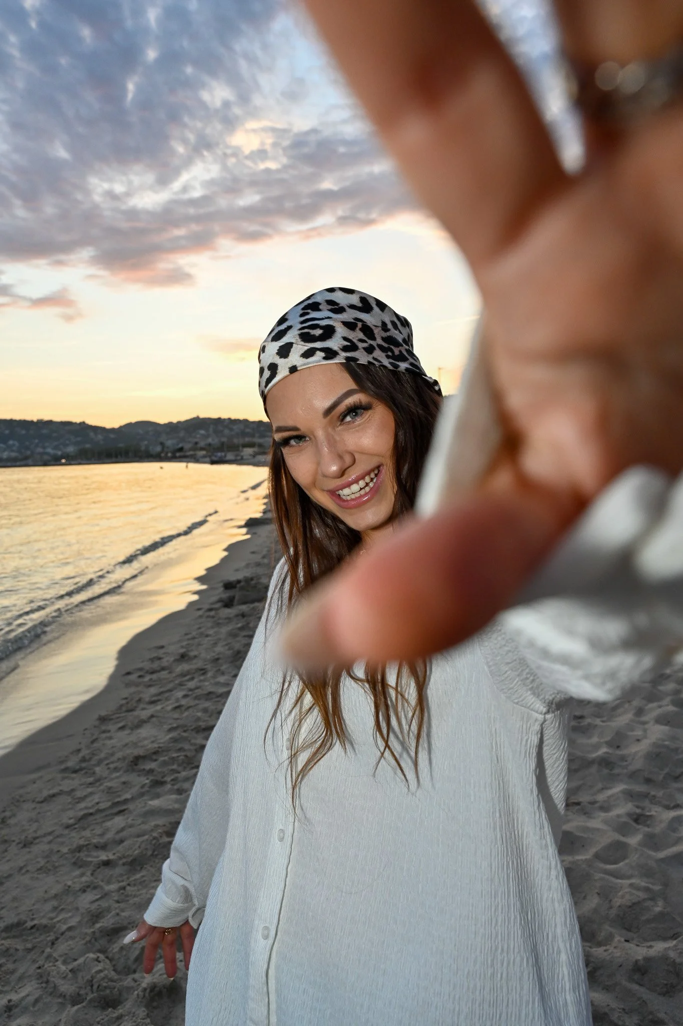 Une femme souriante portant un bandeau à motifs léopard, debout sur la plage au coucher du soleil, une main est approchée vers l'appareil photo, partiellement floue.