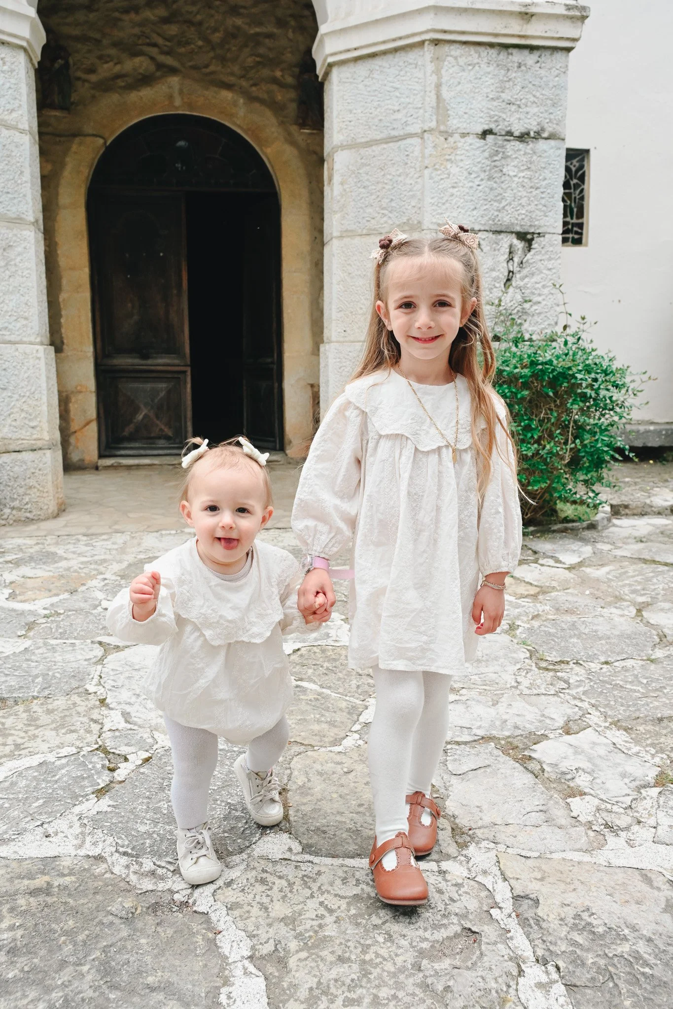 Deux jeunes filles, l'une plus jeune et l'autre un peu plus âgée, portant des robes blanches et des collants, marchent ensemble sur une chaussée en pierre devant une vieille bâtisse en pierre avec une porte en bois.