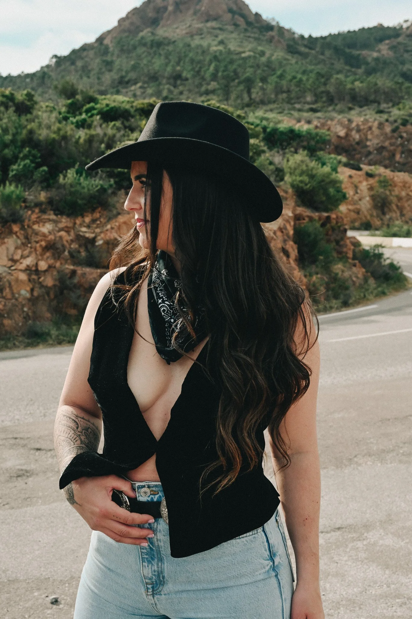 Femme portant un chapeau noir, débardeur noir et bandana noir, posant dans un environnement désertique avec des montagnes en arrière-plan.