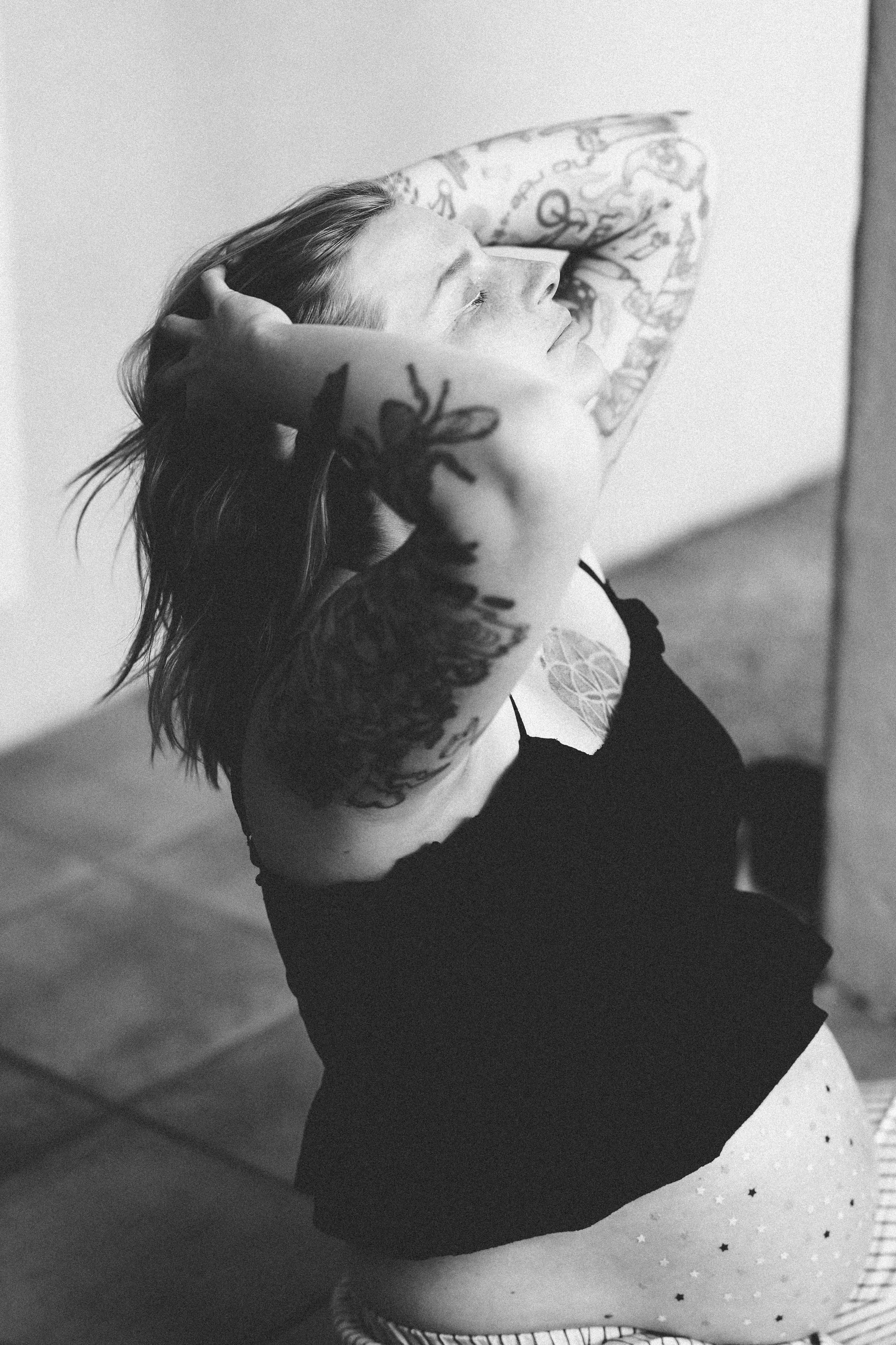 Femme aux cheveux longs et tatouages, portant un haut noir, assise contre un mur, regardant vers le haut, avec des étoiles sur le pantalon.