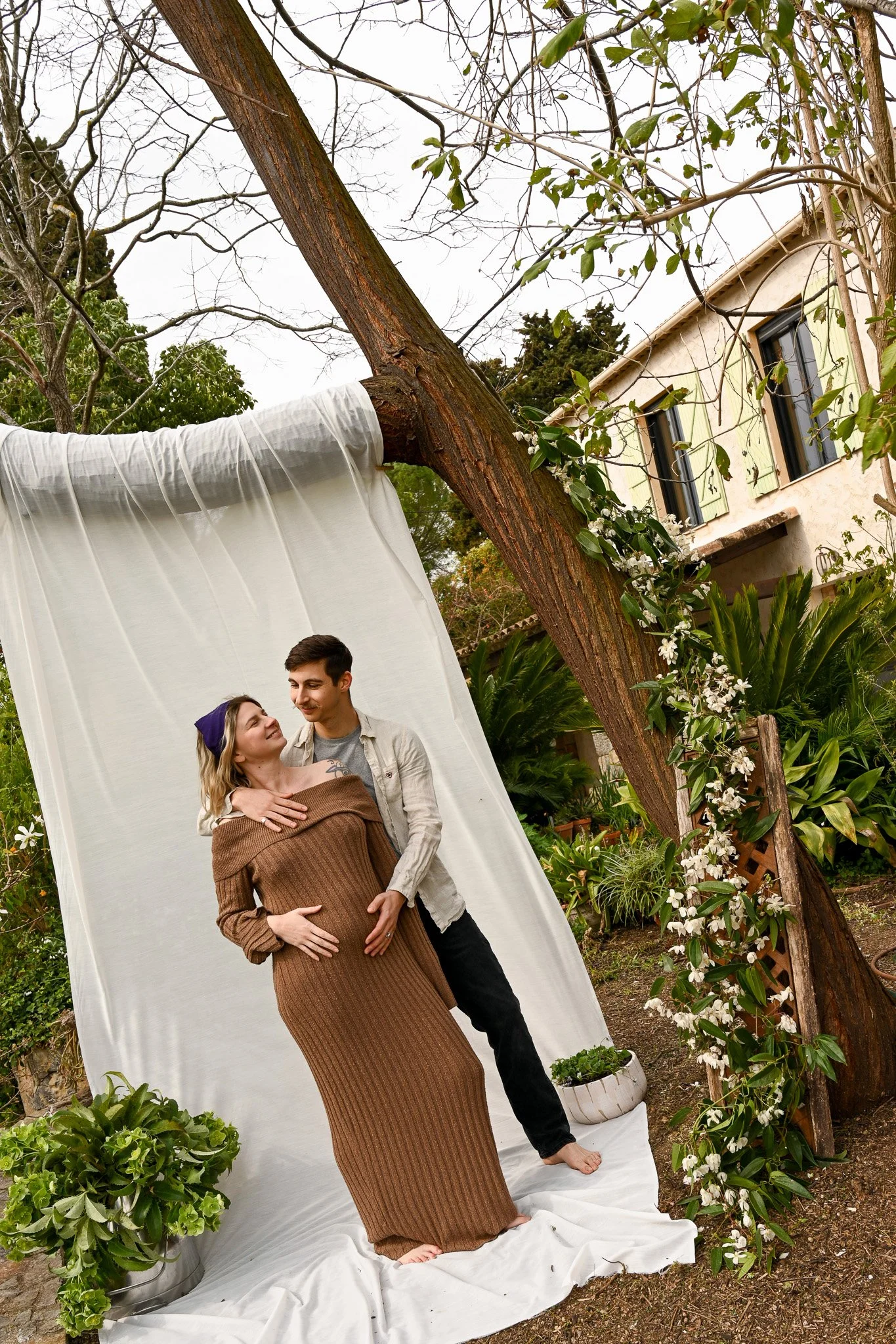 Un couple pose pour une séance photo en extérieur, derrière eux un arbre incliné, un drapé blanc, un jardin avec des plantes en pot et une maison avec des fenêtres à volets verts.