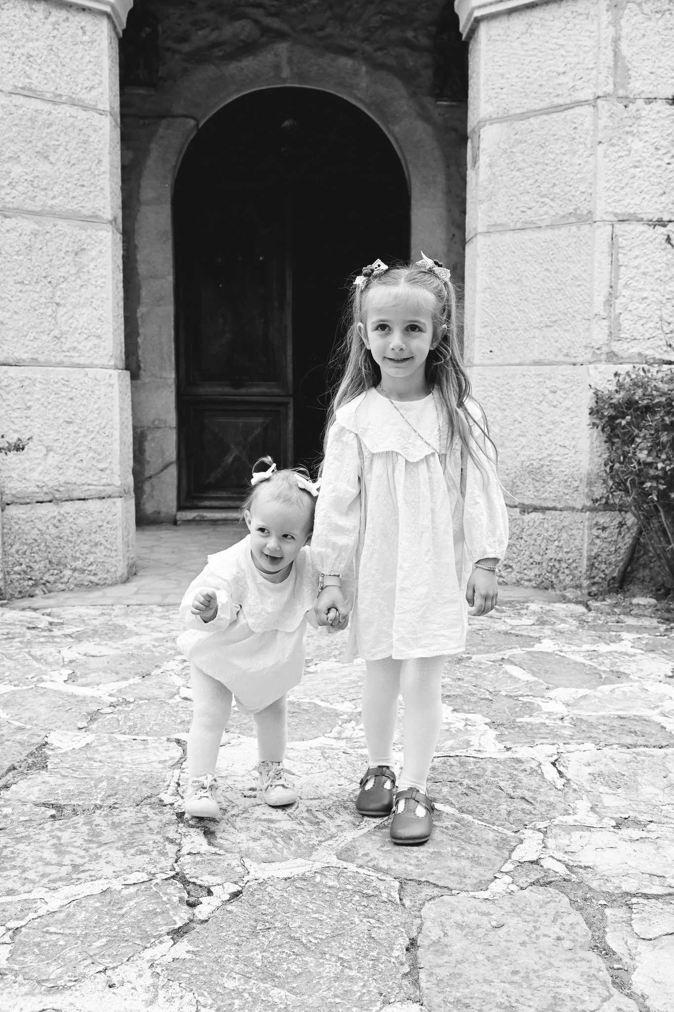 Deux jeunes filles debout sur un pavé en pierre devant une porte en bois encadrée de pierre. La fille plus âgée tient la main de la plus jeune. Elles portent des robes blanches, des collants et des chaussures. La plus jeune a des couettes et la plus 