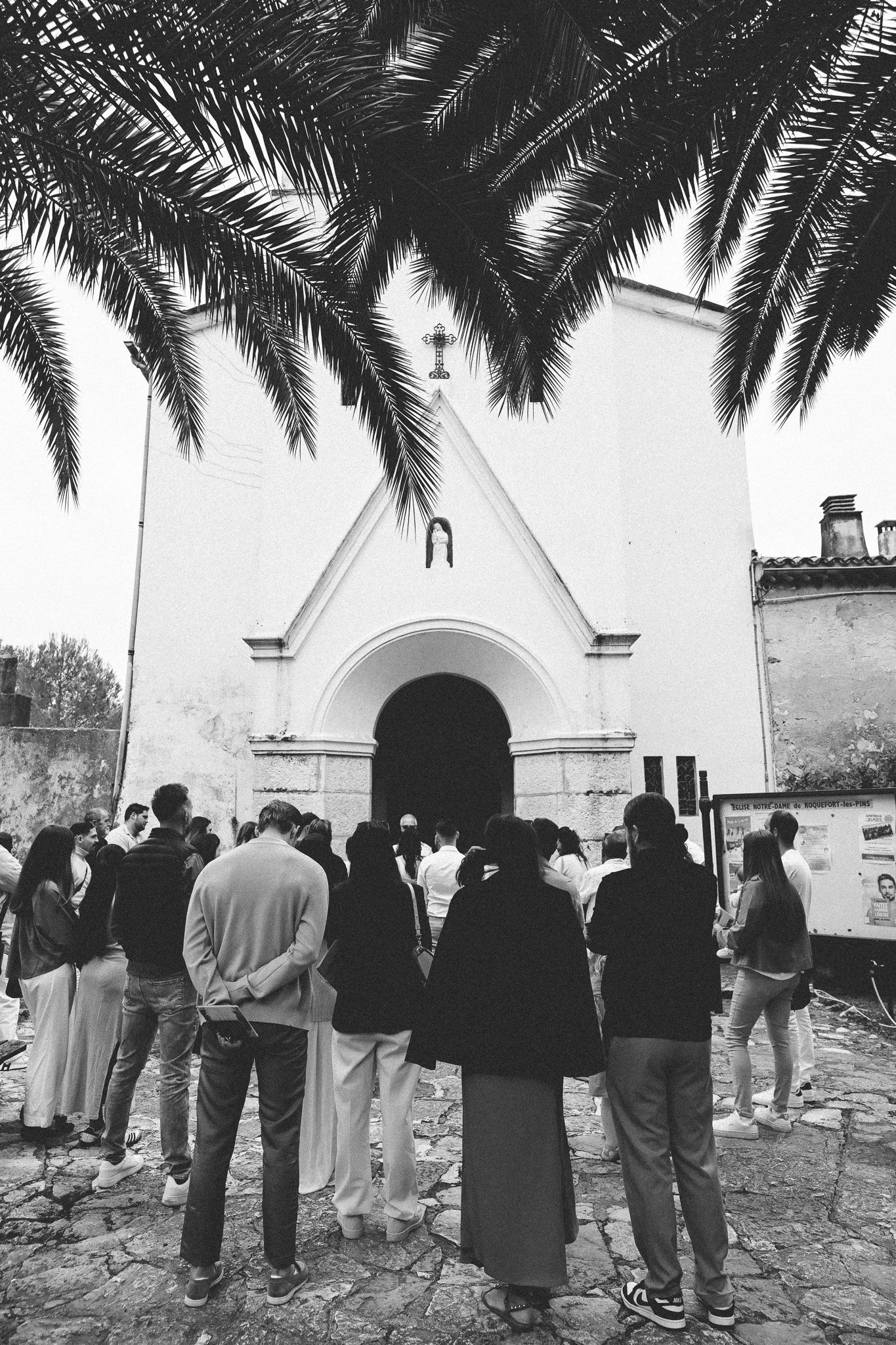 Une foule de personnes se tient devant une église blanche avec une croix à son sommet à Roquefort-les-Pins, sous un arbre à feuilles longues.