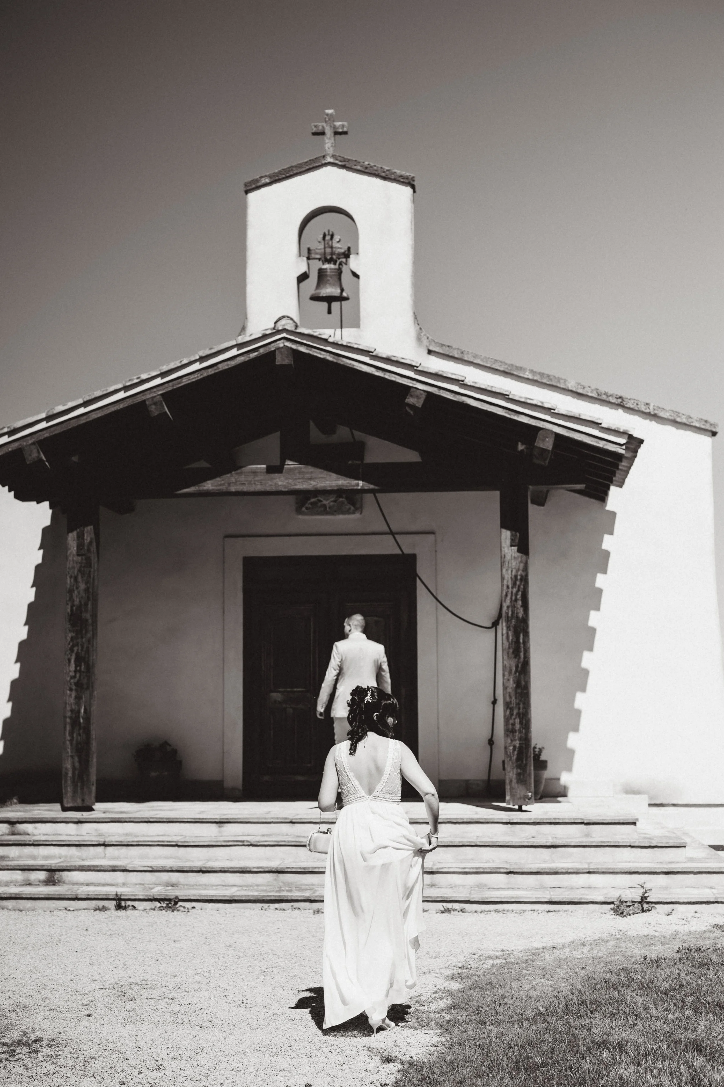 Une femme en robe blanche marche dans la cour de devant d'une petite église avec un homme, elle porte un chapelet dans sa main et un chapeau à fleurs, devant la porte de l'église. La façade de l'église est simple, avec un clocher et une croix au sommet, le tout en noir et blanc.