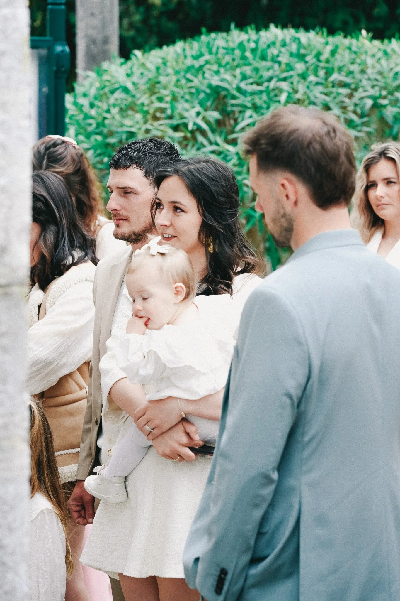 Groupe de personnes assistent à une cérémonie en plein air, y compris un bébé dans les bras d'une femme, le tout dans un environnement naturel avec végétation en arrière-plan.