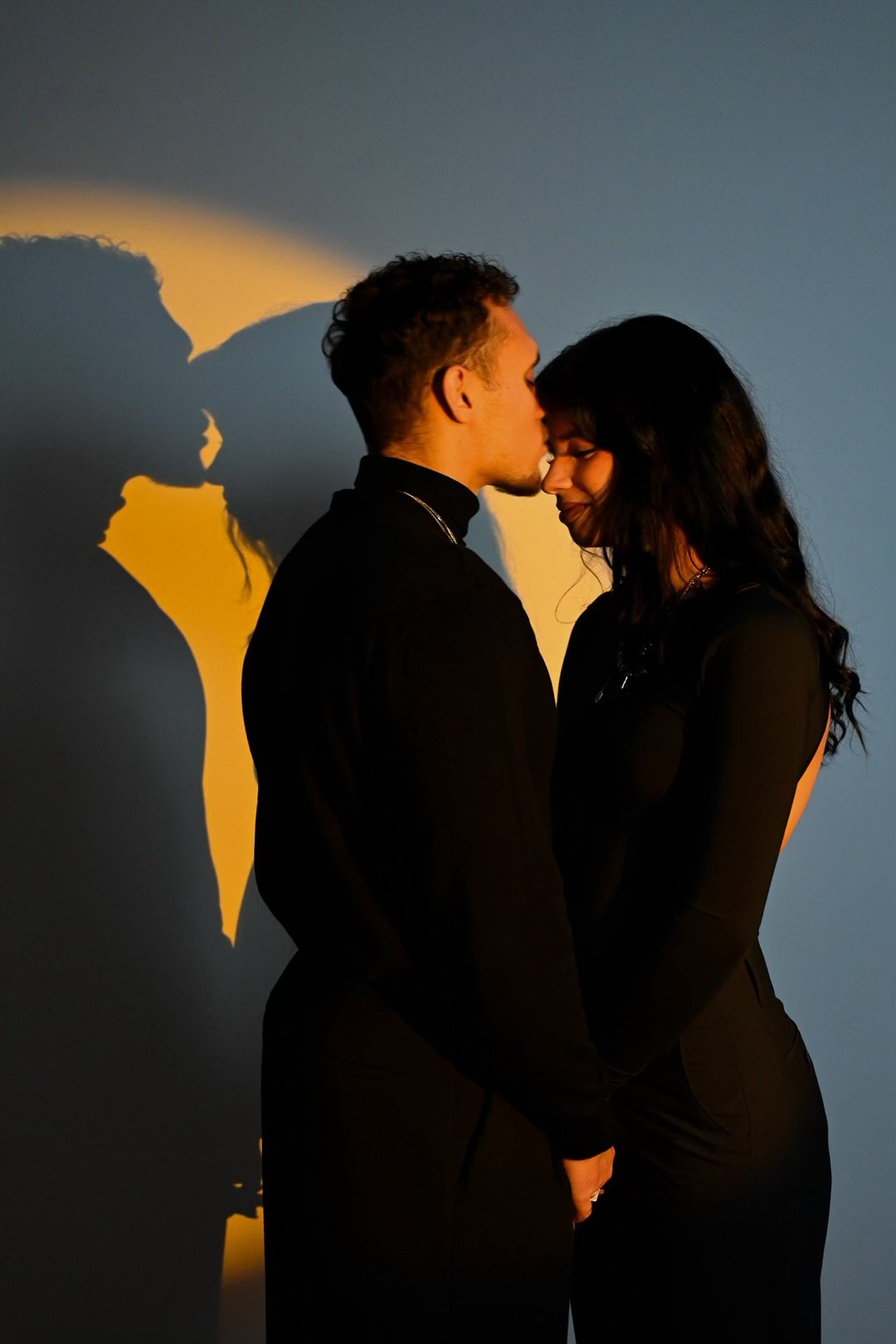 Un jeune homme et une jeune femme se tiennent près d'un mur, leur front en contact, avec un éclairage créant une projection de leur silhouette en forme de cœur.