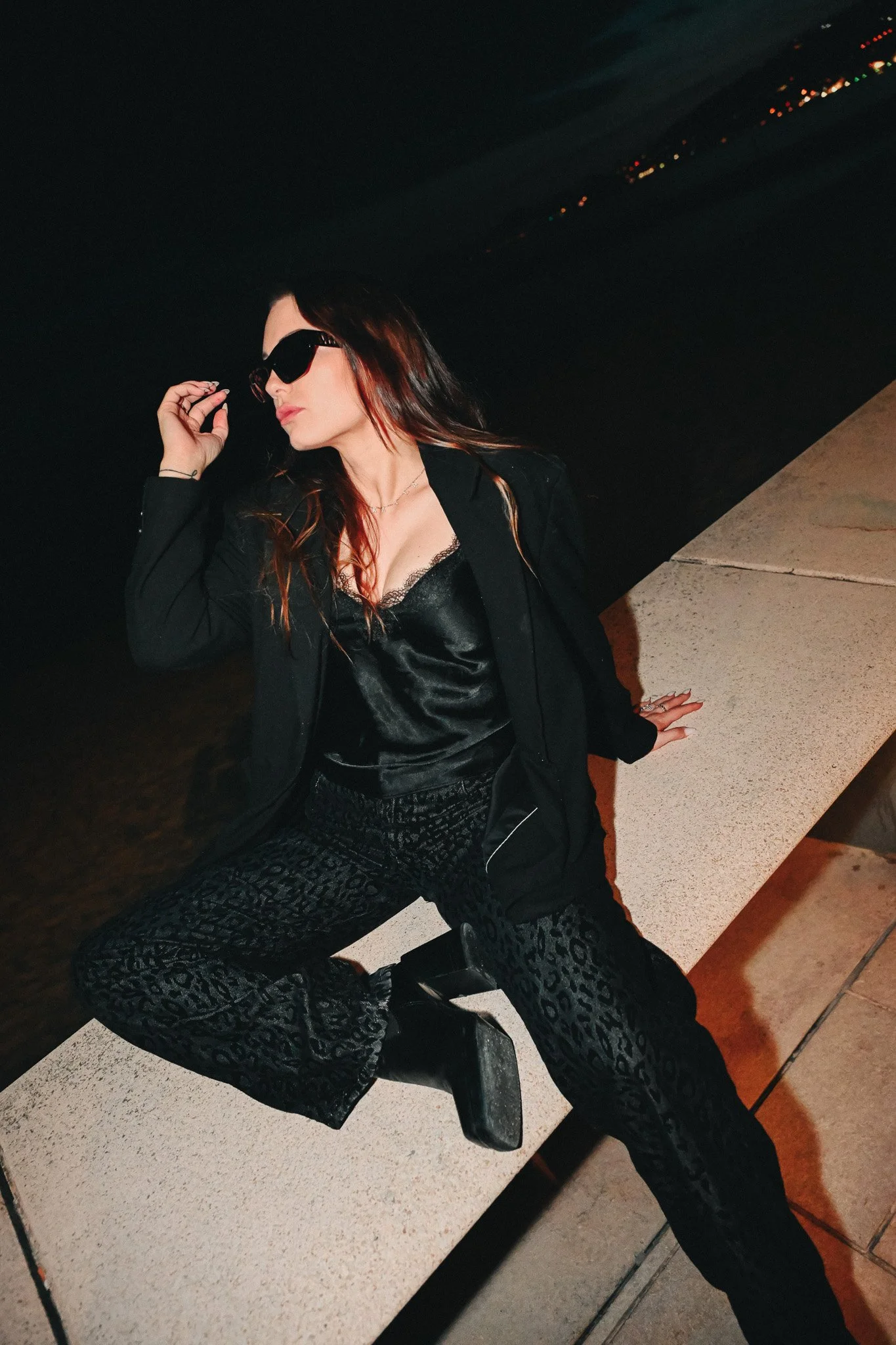 Femme portant des lunettes de soleil noires, vêtue d'une veste noire, assise sur un banc en soirée, avec un fond sombre et des lumières lointaines.
