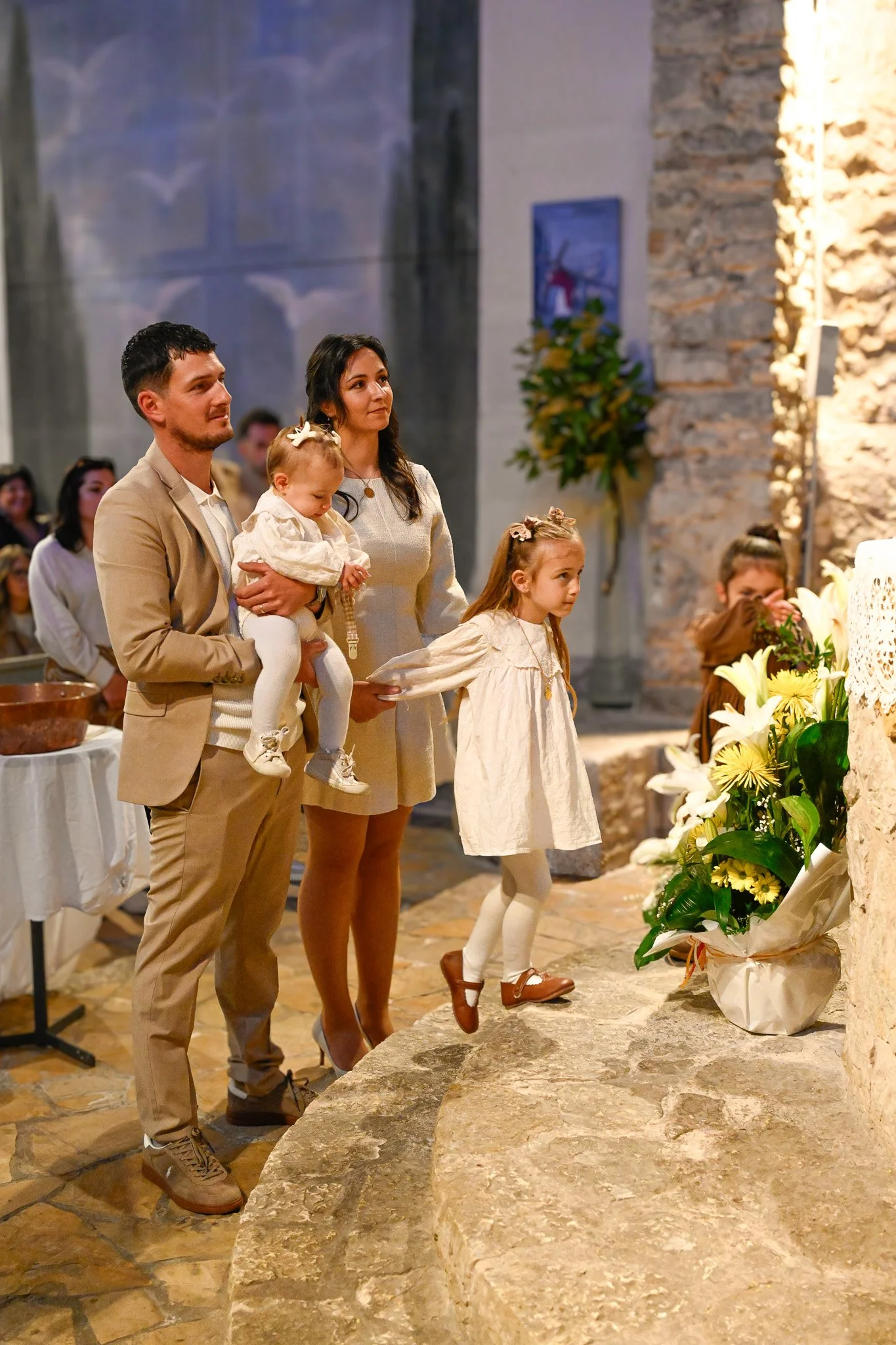 Une famille participe à une cérémonie religieuse dans une église avec un fond de pierres et d'icônes, devant un autel avec des fleurs