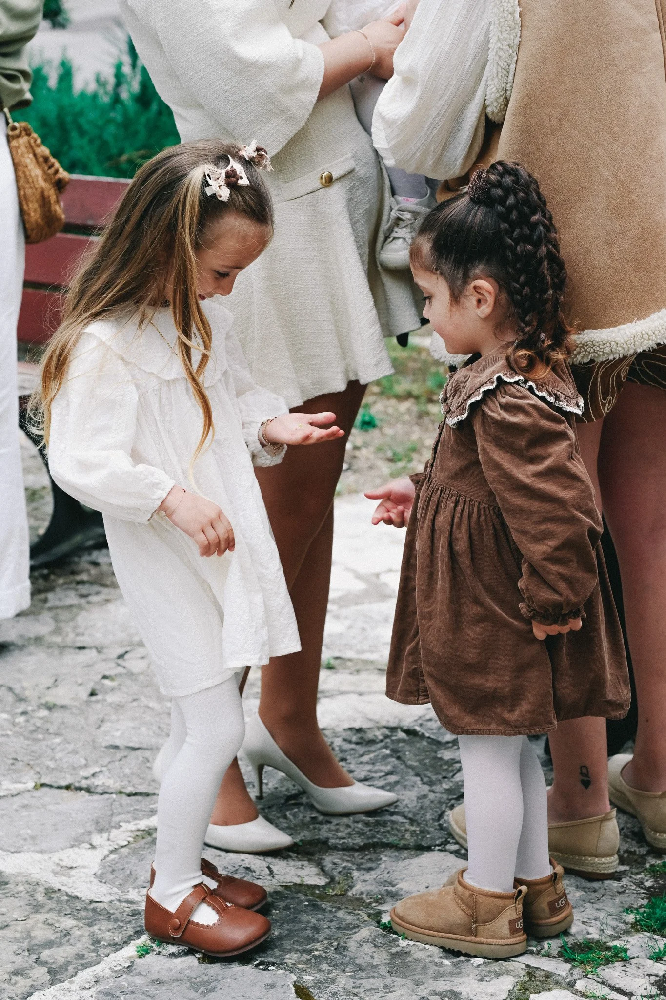 Deux jeunes filles habillées en blanc et en marron, portant des collants blancs et des chaussures marron, debout sur une surface pavée. Elles semblent en train de prendre une photo ou de regarder quelque chose en bas. Plusieurs adultes, en vêtements 