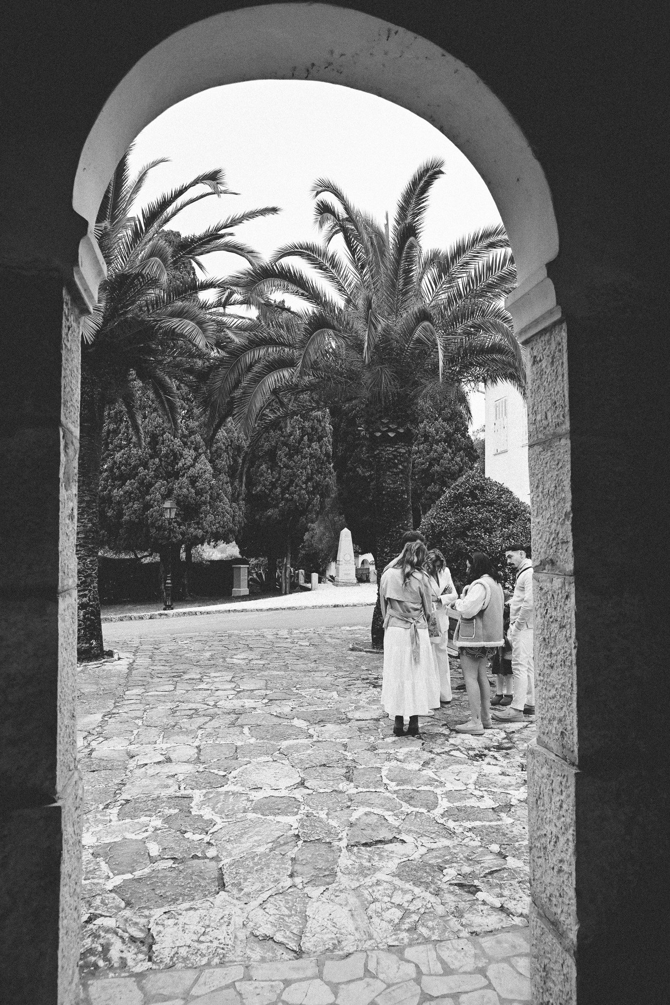 Un groupe de personnes parle dans une cour pavée entourée d'arbres, vus à travers une arche en pierre dans une image en noir et blanc.