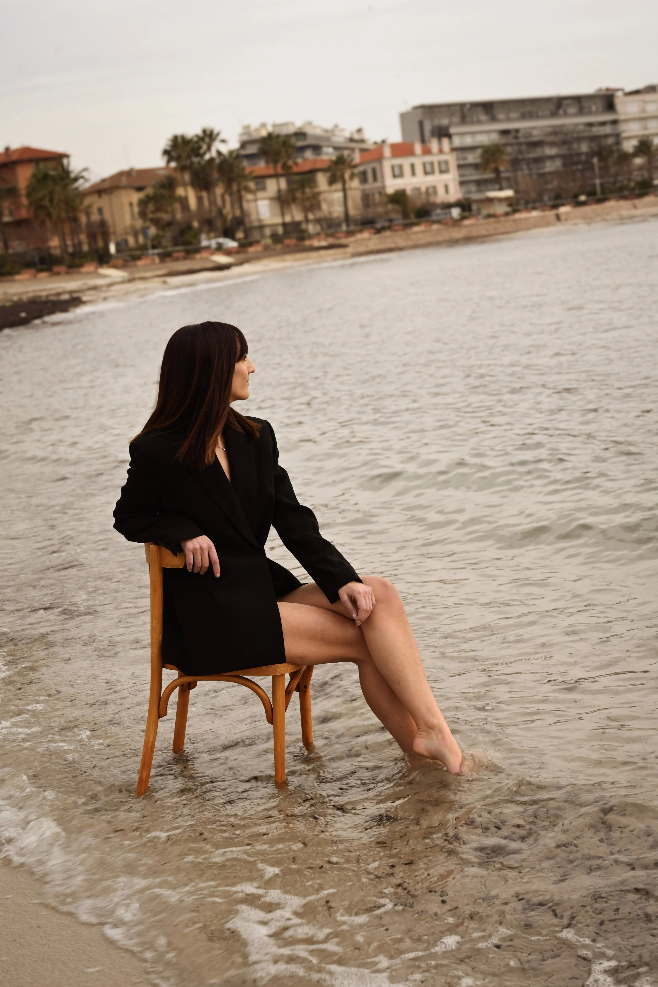 Une femme assise sur une chaise en bois au bord de la mer, avec des bâtiments résidentiels en arrière-plan, ses jambes dans l'eau de mer, portant un blazer noir. L'ambiance est calme et la mer calme.
