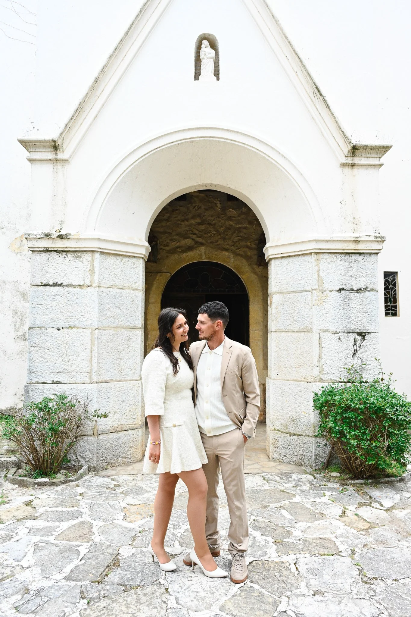 Un couple habillé élégamment se tenant près d'une entrée d'église en pierre, avec un homme en costume beige et une femme en robe blanche, souriant et se regardant affectueusement.