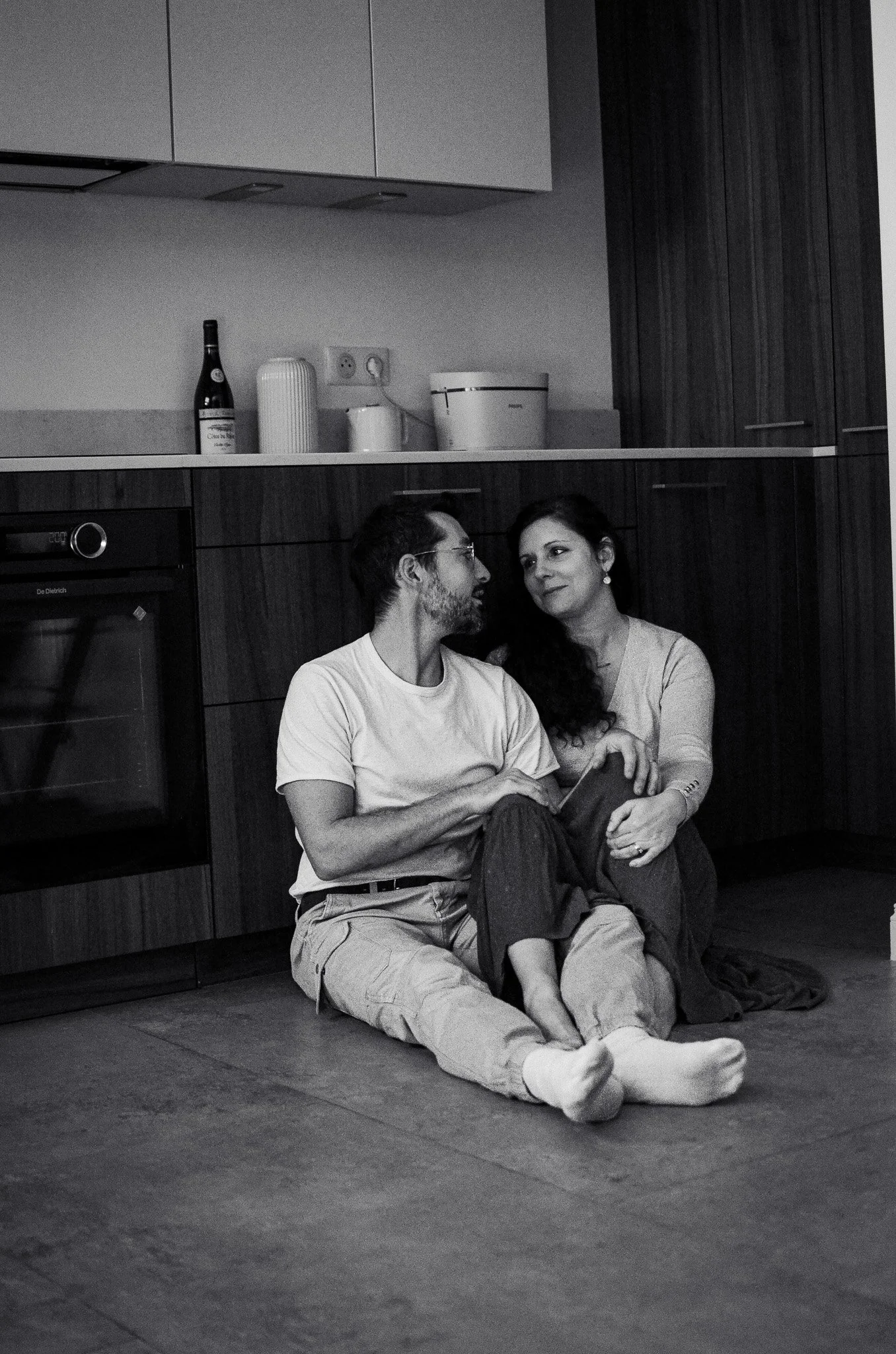 Un homme et une femme assis par terre dans une cuisine, la femme regarde l'homme avec un sourire, ils sont proches, l'homme tient un journal ou une tablette.