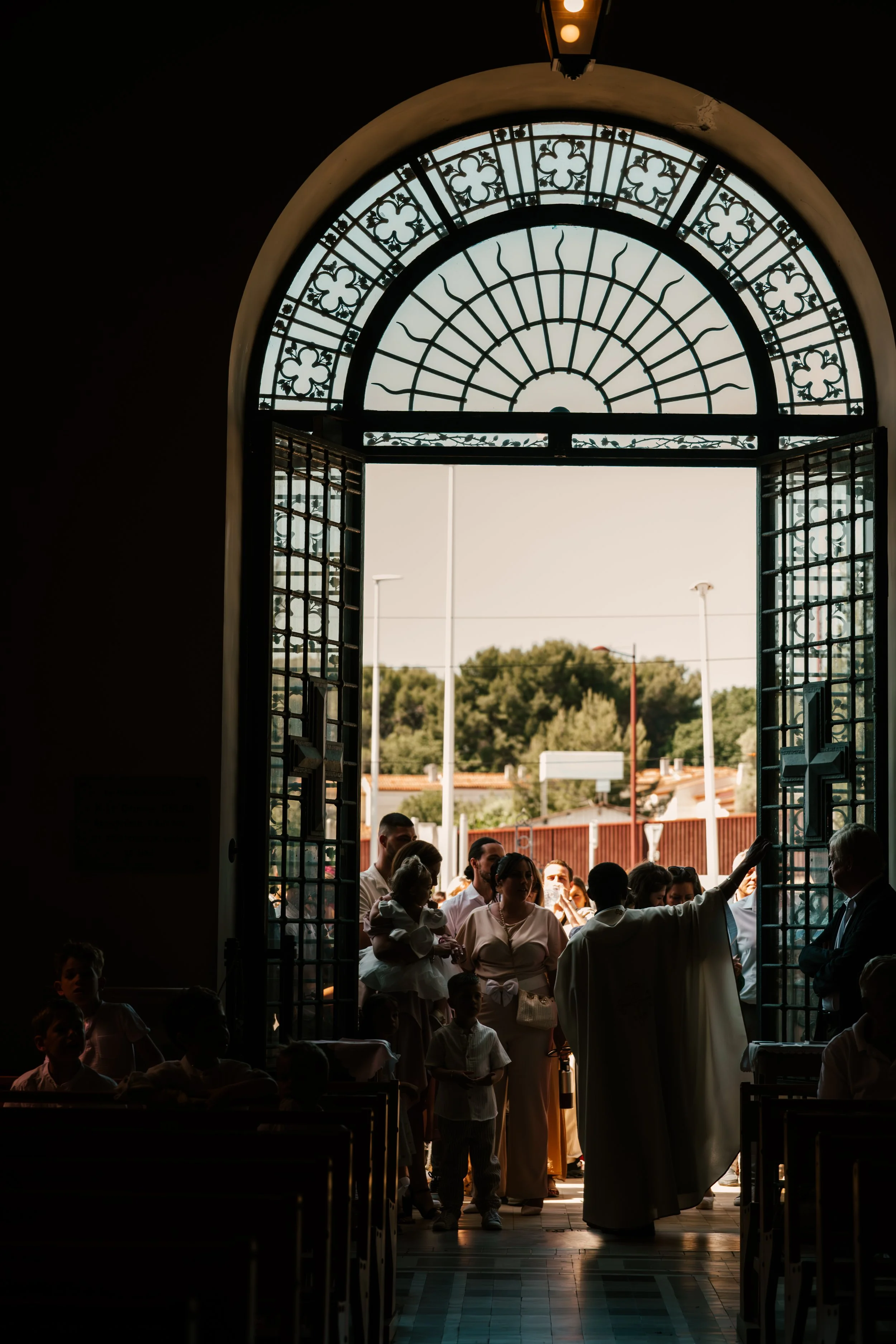 Une vue intérieure d'une église avec des portes ouvertes laissant passer la lumière du soleil sur un groupe de personnes rassemblées. La structure de l'entrée est ornée de vitraux en forme de trèfle.