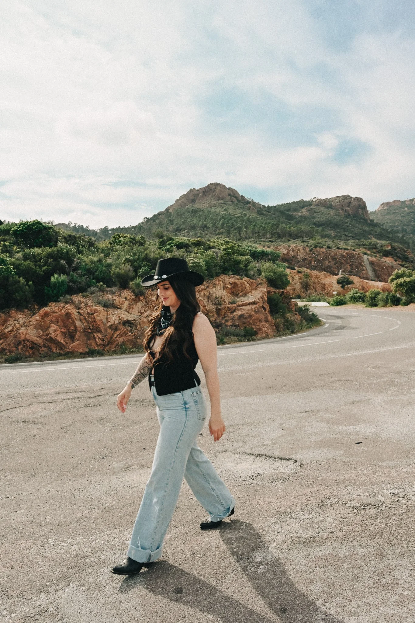 Femme avec long cheveux foncés portant un chapeau noir, un foulard noir à pois et un haut noir, marchant sur une route de campagne entourée de rochers et de végétation, avec des montagnes en arrière-plan et un ciel partiellement nuageux.