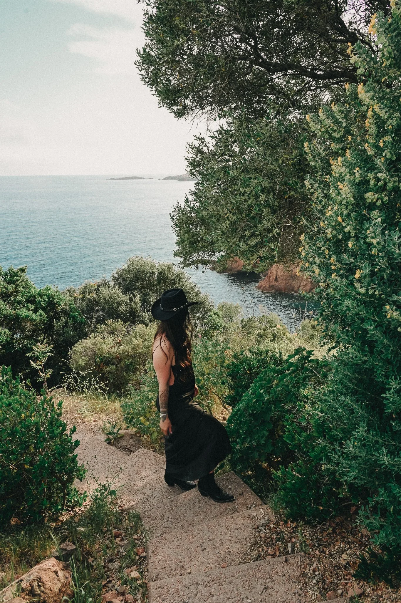 Une femme portant un chapeau noir et une robe noire marche en descendant des escaliers en pierre, entourée de végétation verte, avec une vue sur la mer et le ciel clair en arrière-plan.