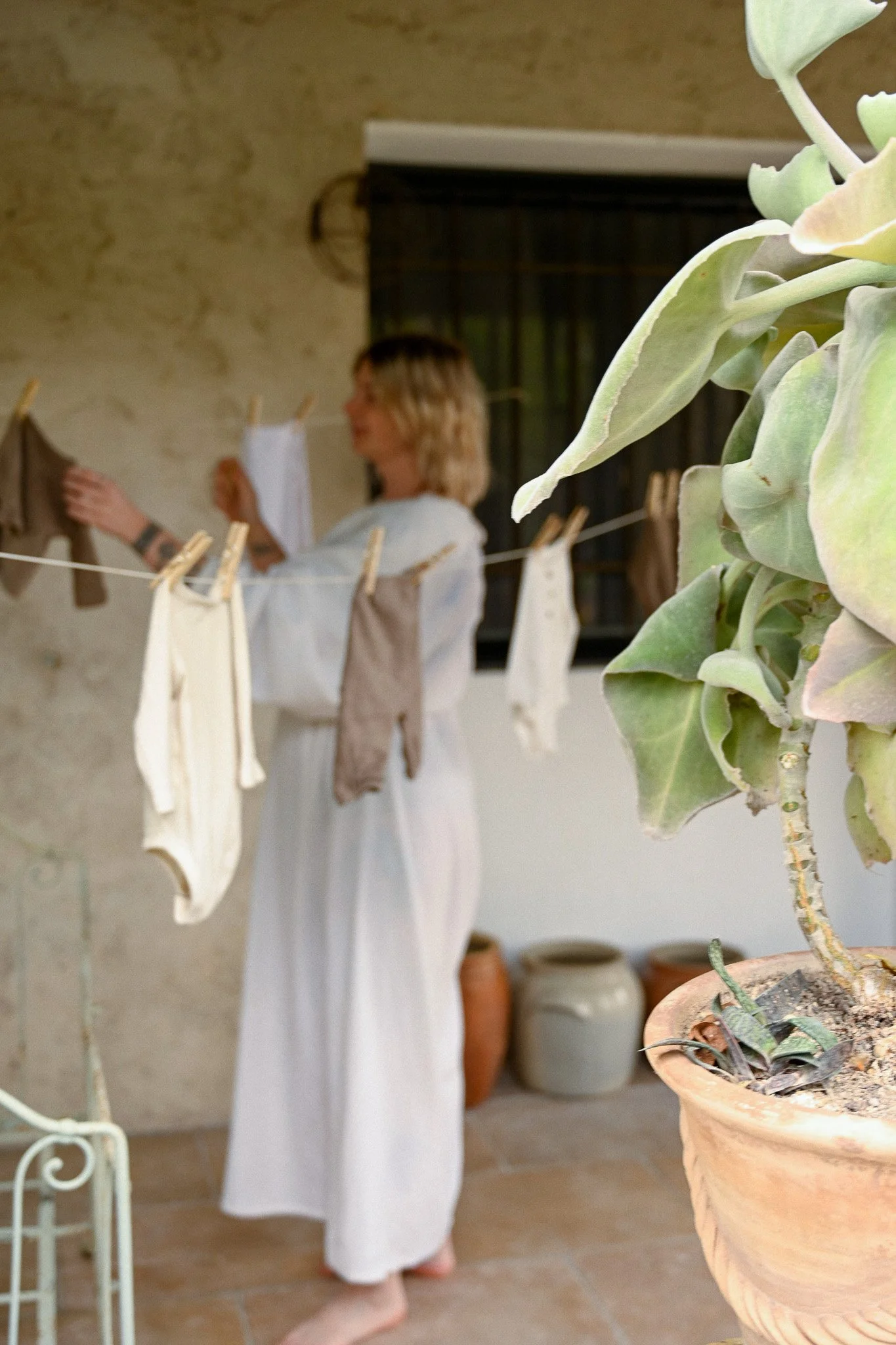 Une femme en robe blanche suspend des vêtements ou du linge propre sur une corde à linge avec des pinces, dans un intérieur avec un mur en plâtre, et des pots en terre cuite au sol. Un gros plant de Plante succulente est en premier plan, à droite.