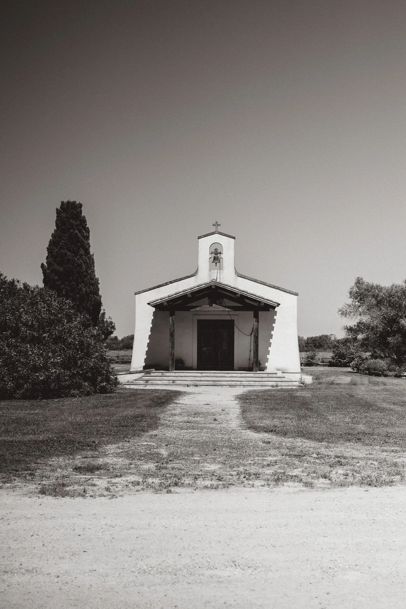 Une petite église blanche avec un clocher abritant une cloche, entourée d'arbres, avec un chemin de terre devant, en noir et blanc.