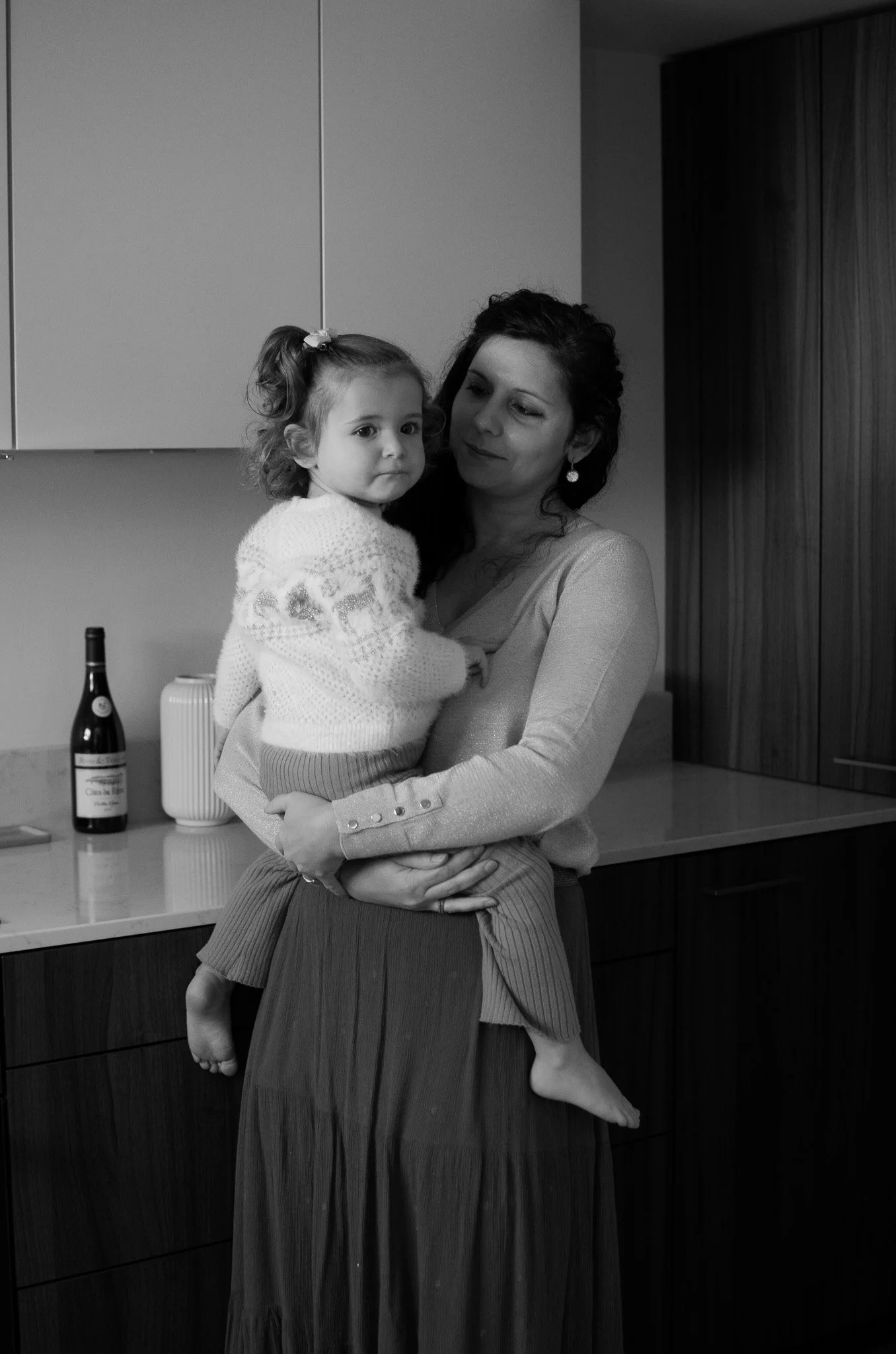 Une femme tient une jeune fille dans ses bras dans une cuisine. La jeune fille, assise sur la taille de la femme, a les jambes pendantes et porte un pull blanc et une jupe longue. La cuisine a un plan de travail avec une bouteille de vin, une boîte e