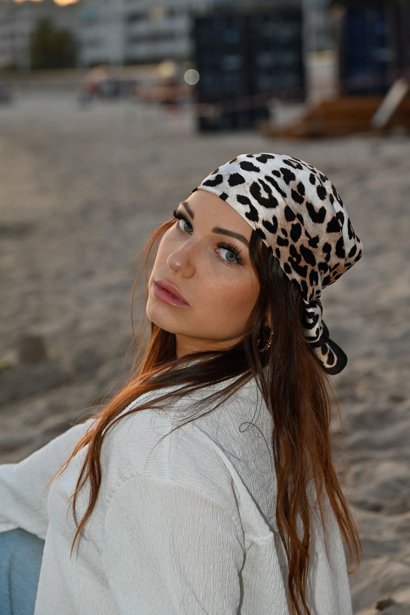Une jeune femme portant un foulard à motifs léopard et un t-shirt blanc, posant à l'extérieur près d'une zone de construction ou de stockage avec des bâtiments en arrière-plan.
