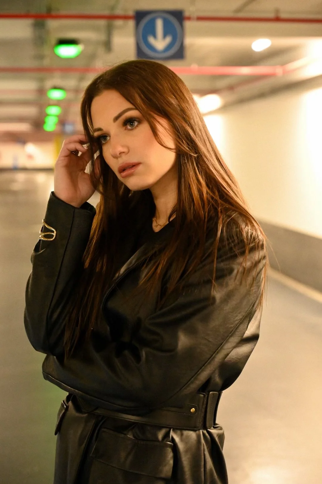 Jeune femme aux cheveux roux, portant une veste en cuir noire, dans un parking souterrain avec un panneau bleu indiquant la direction vers le bas.