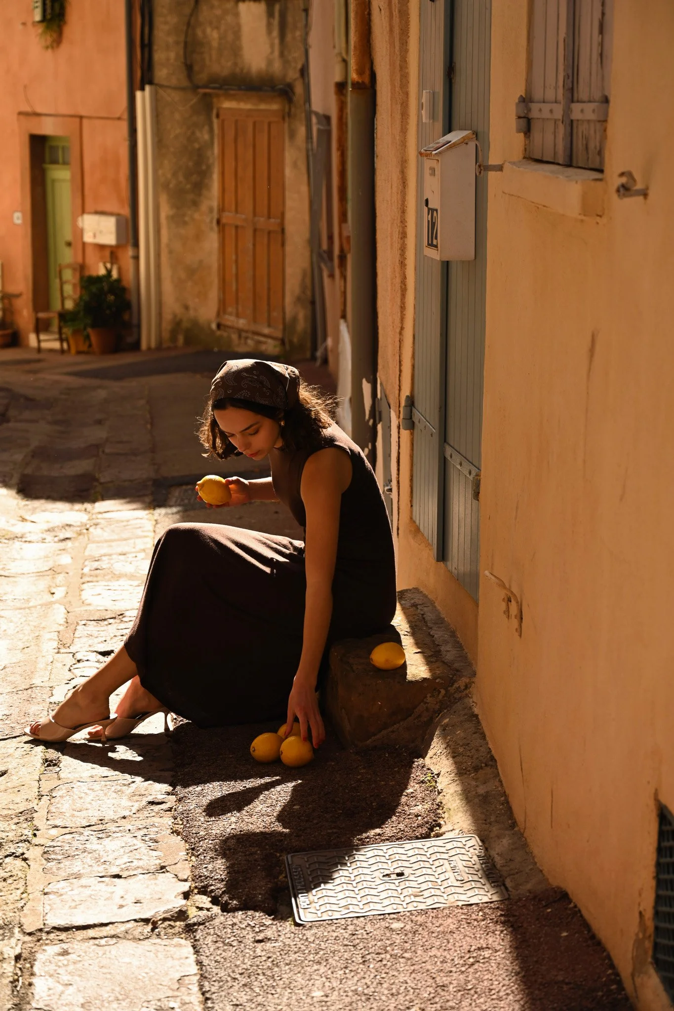 Une femme assise sur un rebord de pierre dans une rue ensoleillée, tenant un citron dans sa main, avec d'autres citrons à ses pieds, à côté d'une maison avec des volets en bois et une porte.